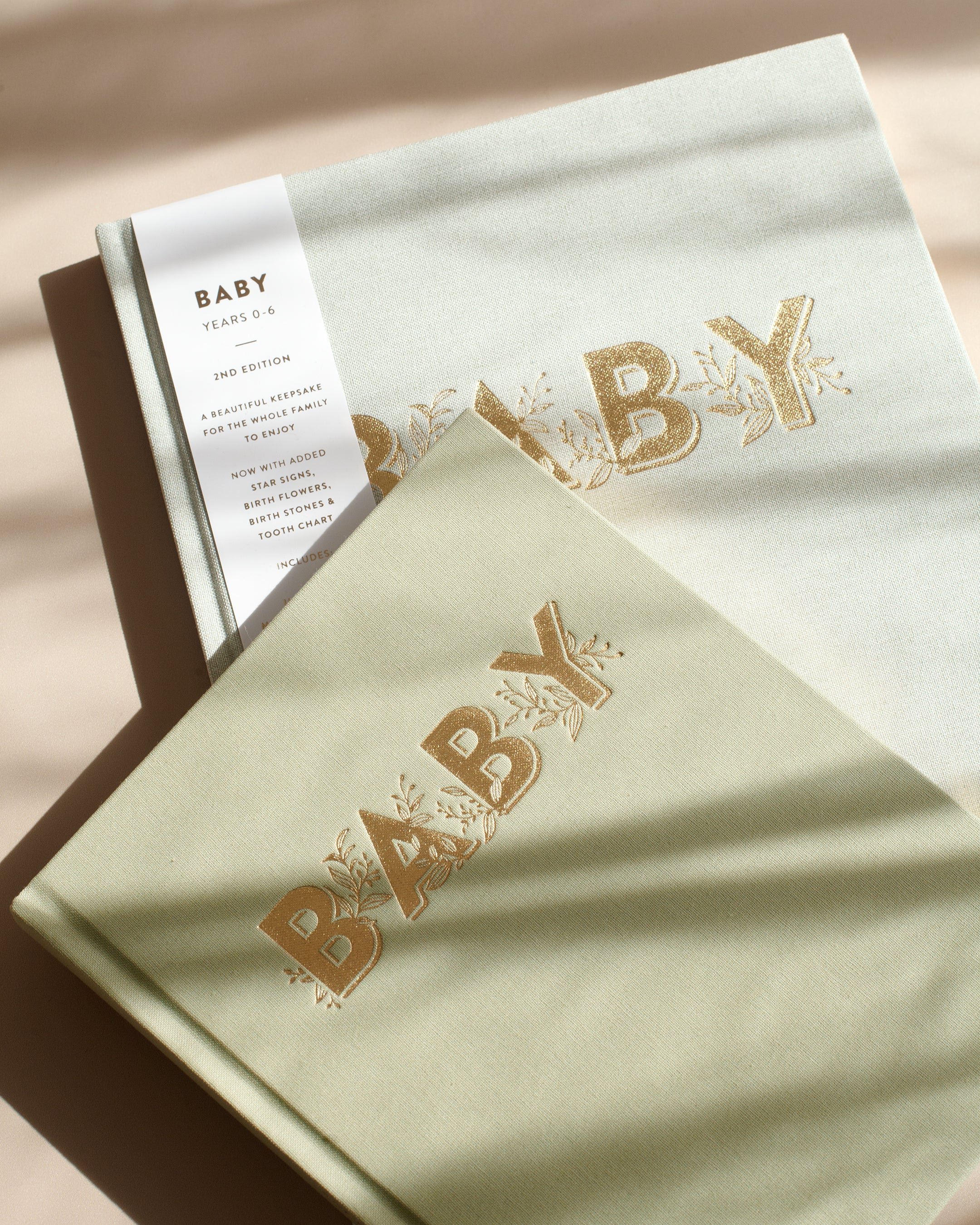 Two FOX & FALLOW Baby Book Pistachios with neutral-colored covers lie on a beige surface. Both covers feature the word "BABY" in gold capitals with floral accents. One book, labeled as the 2nd edition, includes added pages and sections for baby's first five years, making it a perfect family keepsake.