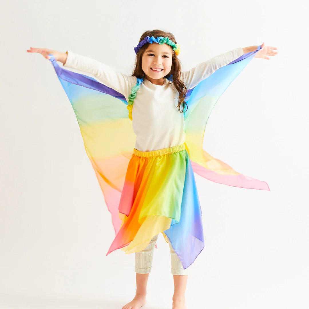 A young girl stands smiling with her arms outstretched, embracing the magic of dress-up. She is wearing Rainbow Wings from SARAH'S SILKS, complete with a matching colorful skirt. A blue and green floral headband adorns her head against a plain white background.