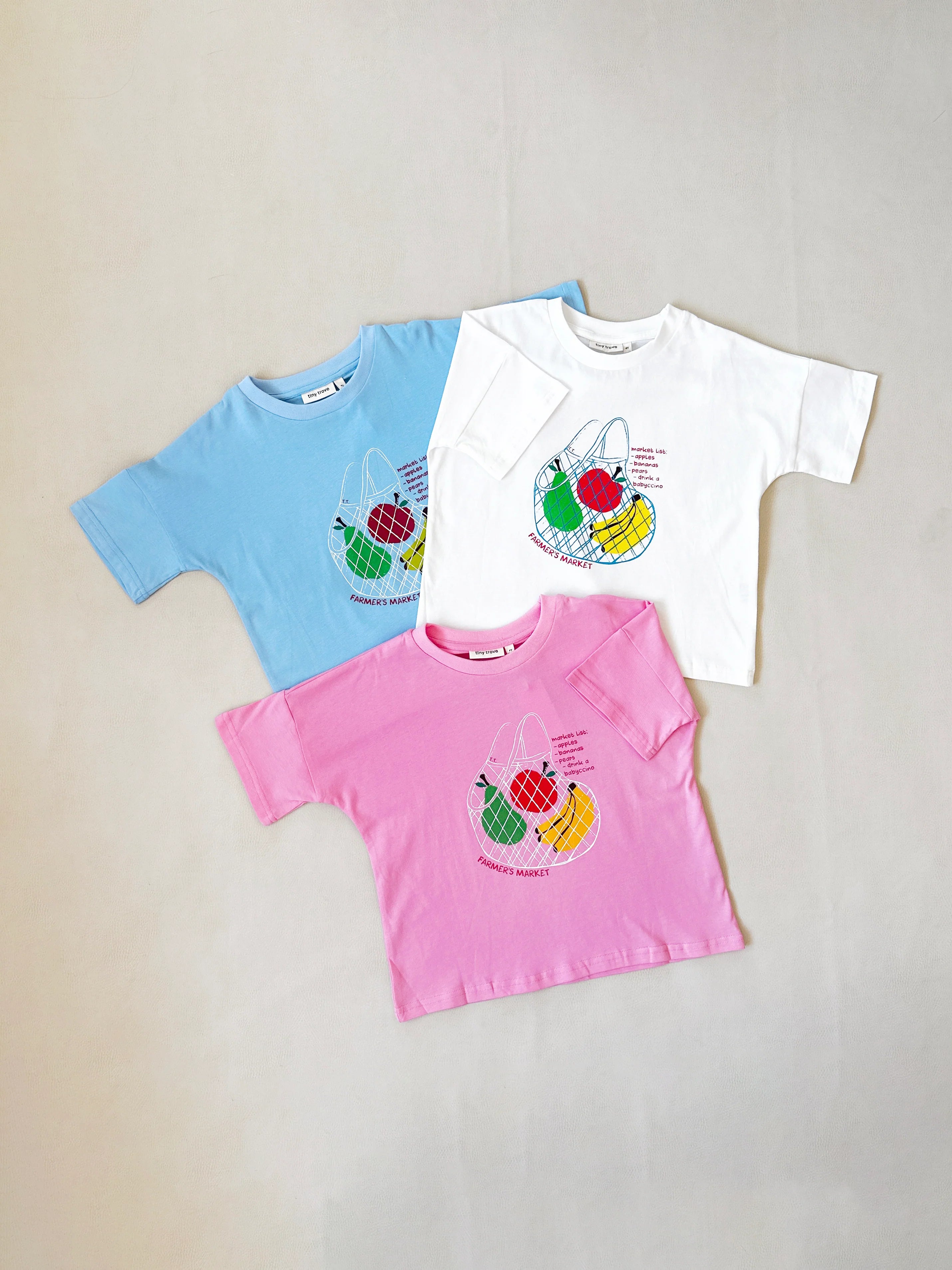 Three TINY TROVE "Farmer's Market Relaxed Tees" in blue, white, and pink are displayed. Made from soft cotton, each features a vibrant basket of vegetables graphic and the phrase "Happy Market.