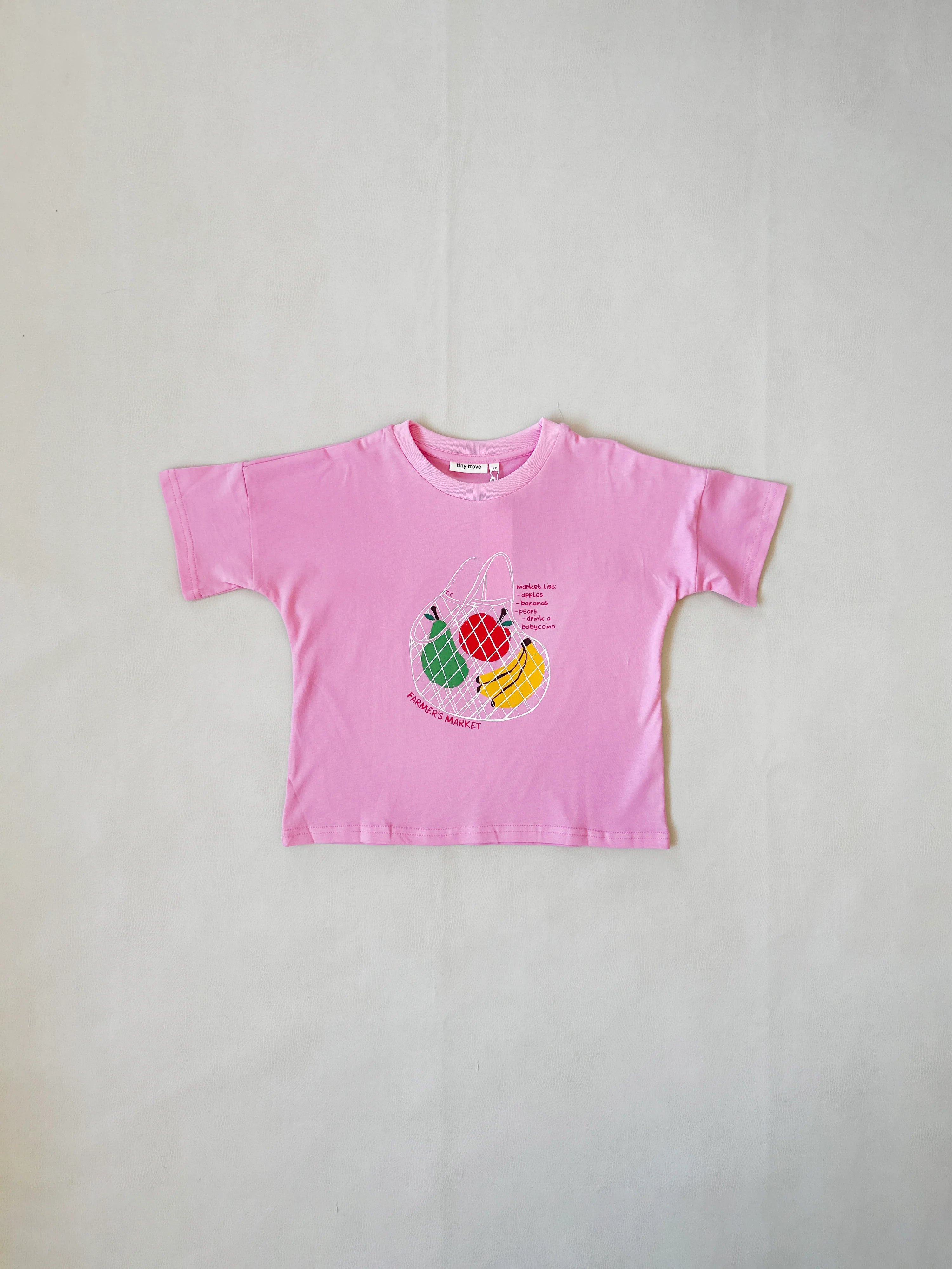 The Tiny Trove ~ Farmer's Market Relaxed Tee Bubblegum showcases fruit graphics like strawberry, kiwi, and mango with skateboards. Made from soft cotton, this pink short-sleeved TINY TROVE tee is displayed flat against a light background.