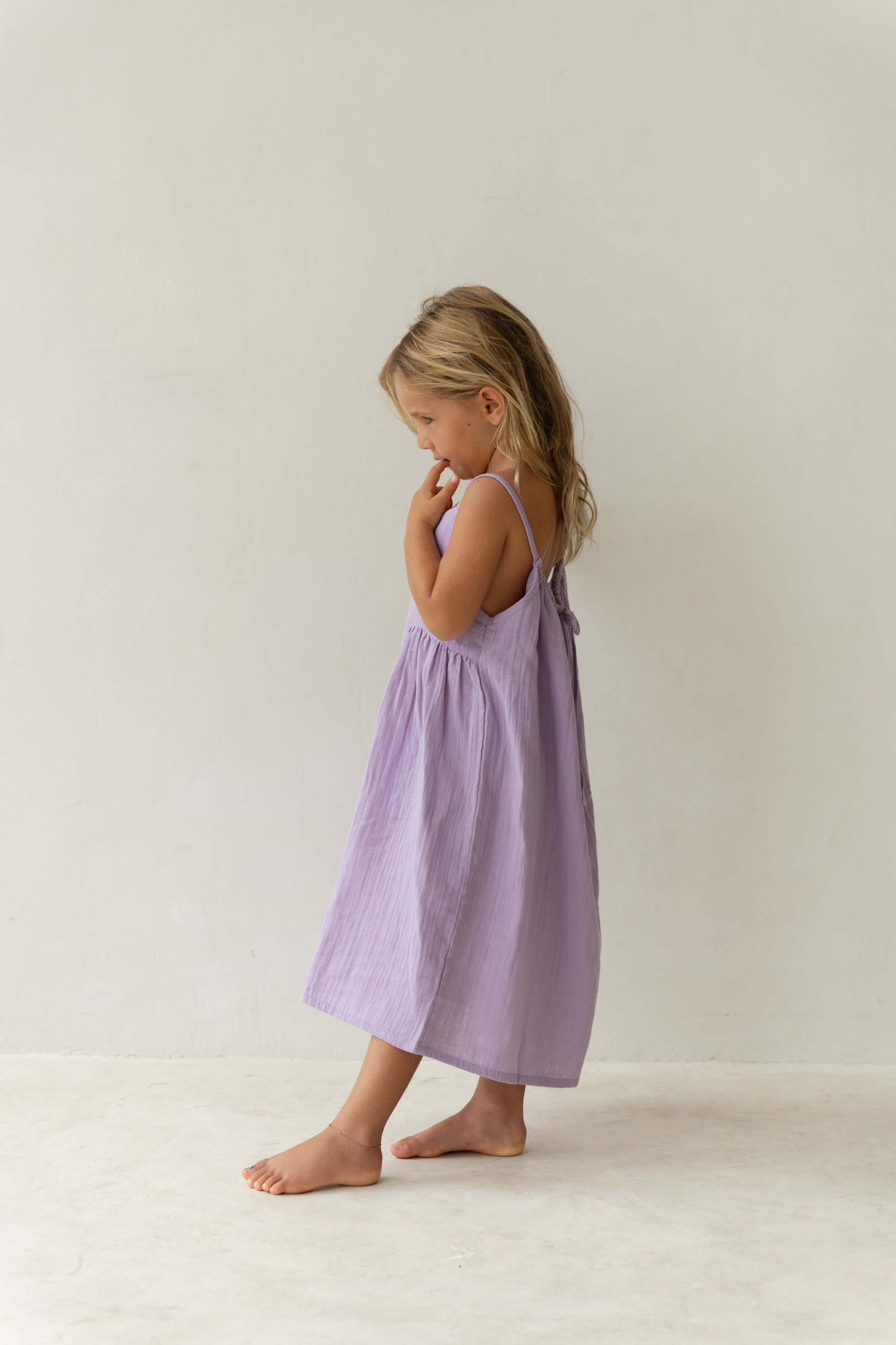 A young girl, standing barefoot against a light background, wears the ILLOURA THE LABEL Field Dress Lilac - Size 1. Her long, light brown hair falls over her shoulders as she gazes downward thoughtfully.