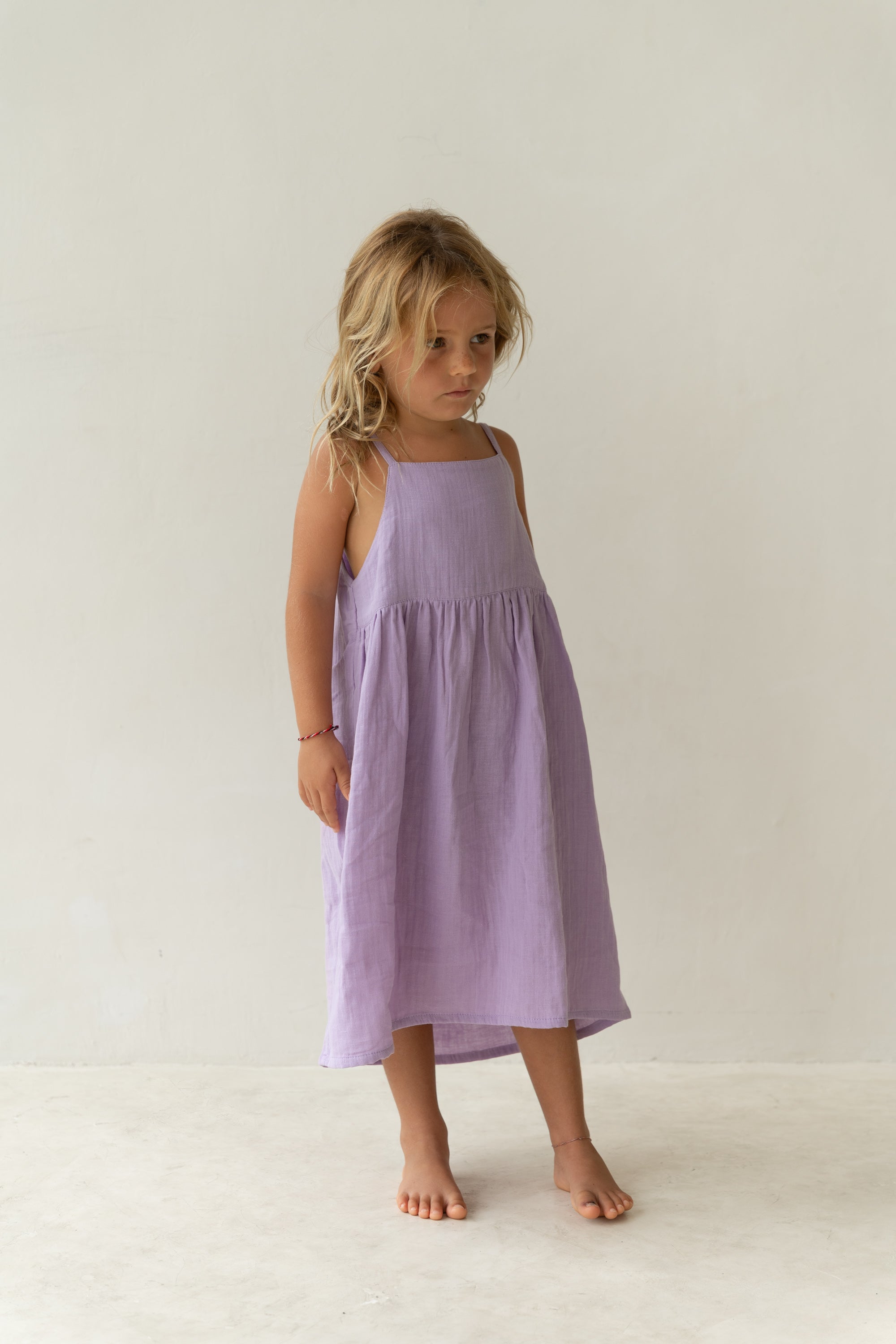 A young child with blonde hair stands barefoot on a light-colored floor against a plain off-white background, wearing the Field Dress Lilac by ILLOURA THE LABEL. The sleeveless, light purple cotton voile midi dress gently drapes as the child gazes slightly downward with a thoughtful expression.