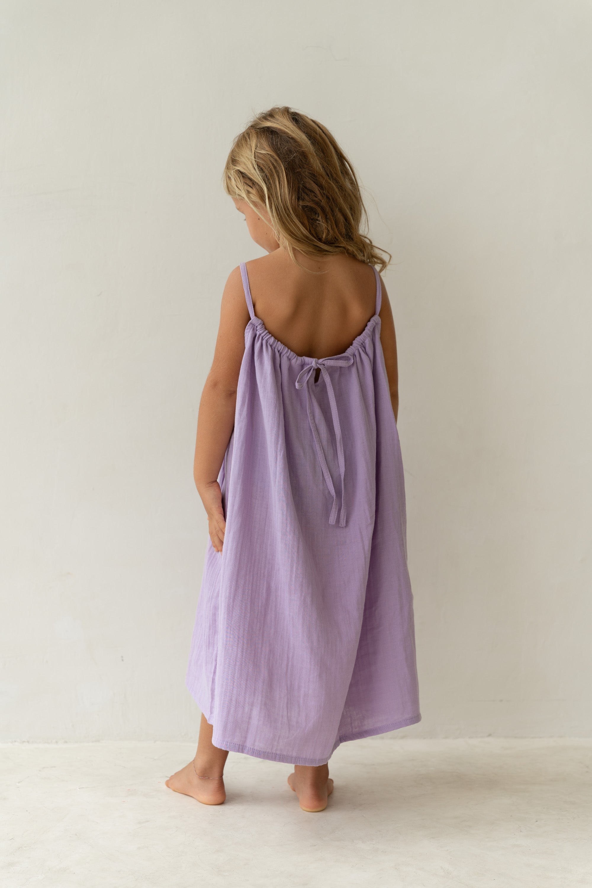 A barefoot young girl with long blond hair stands on a light floor, slightly turned away. She wears the ILLOURA THE LABEL's Field Dress in Lilac (Size 1), a loose lavender midi with thin straps and a tie back, made of soft cotton voile. The background is a plain light wall.