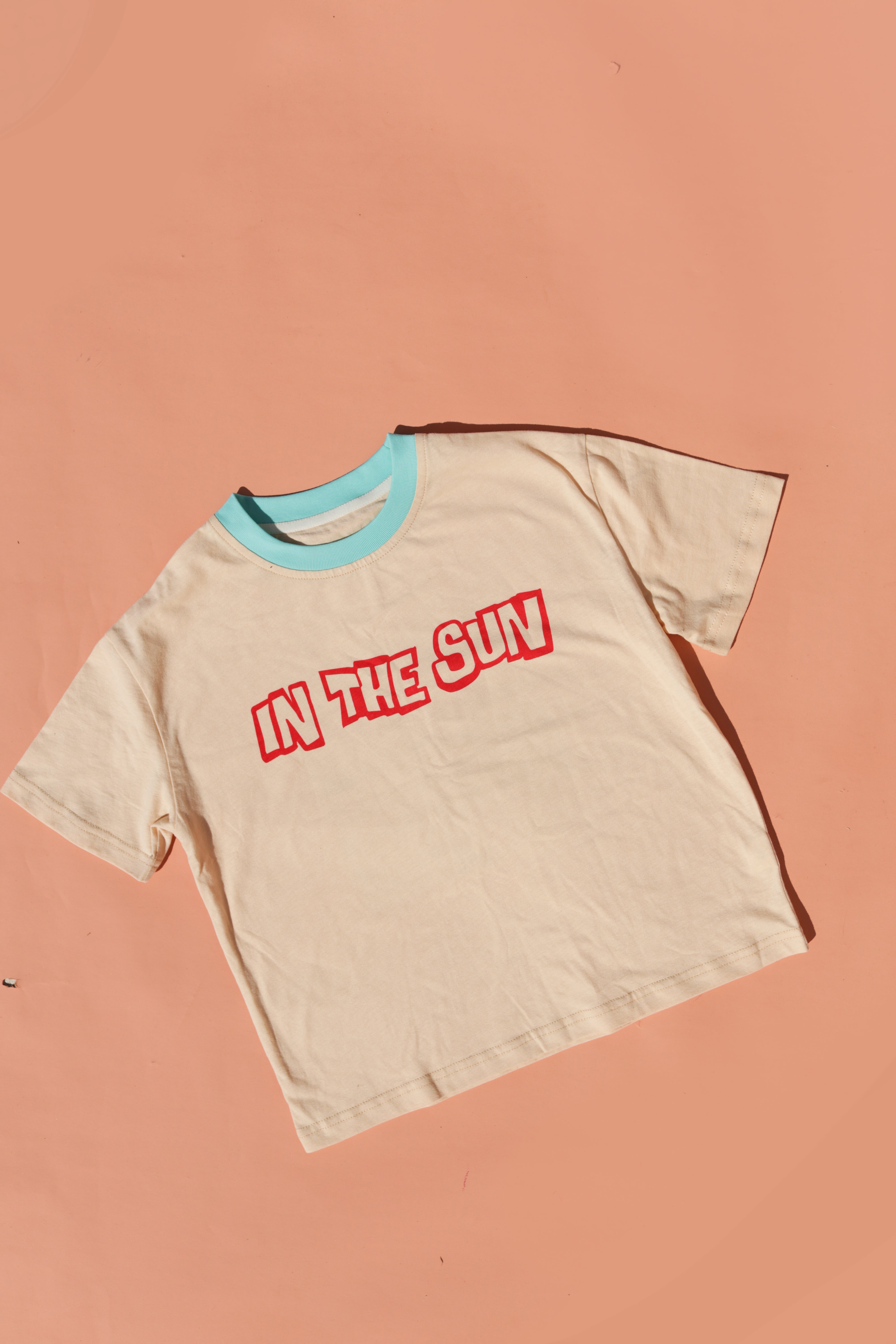 The SMALL SWIM CLUB's In the Sun Tee Cream lies flat on a peach background. Made from soft cotton, this beige surfer tee features a turquoise collar and bold red graphic text that reads "IN THE SUN" at the center.