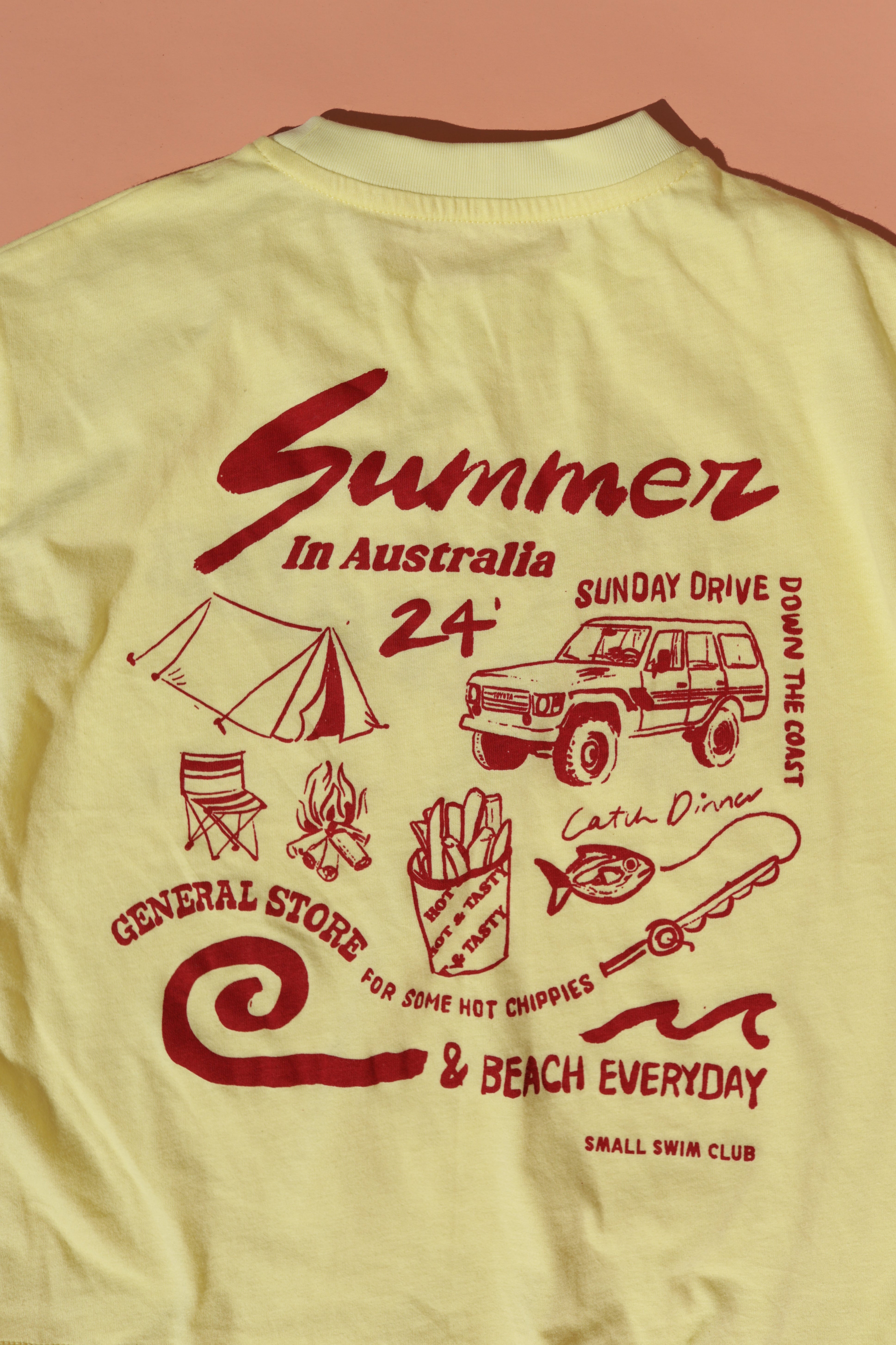 The "Summer in Australia Tee Lemon" by SMALL SWIM CLUB features a yellow base with maroon text and red graphics, depicting camping gear, a car, a campfire, and fries. Phrases include "Summer in Australia 24'," "Sunday Drive," "Catch Dinner," "General Store," and "& Beach Everyday." Crafted from 100% cotton.