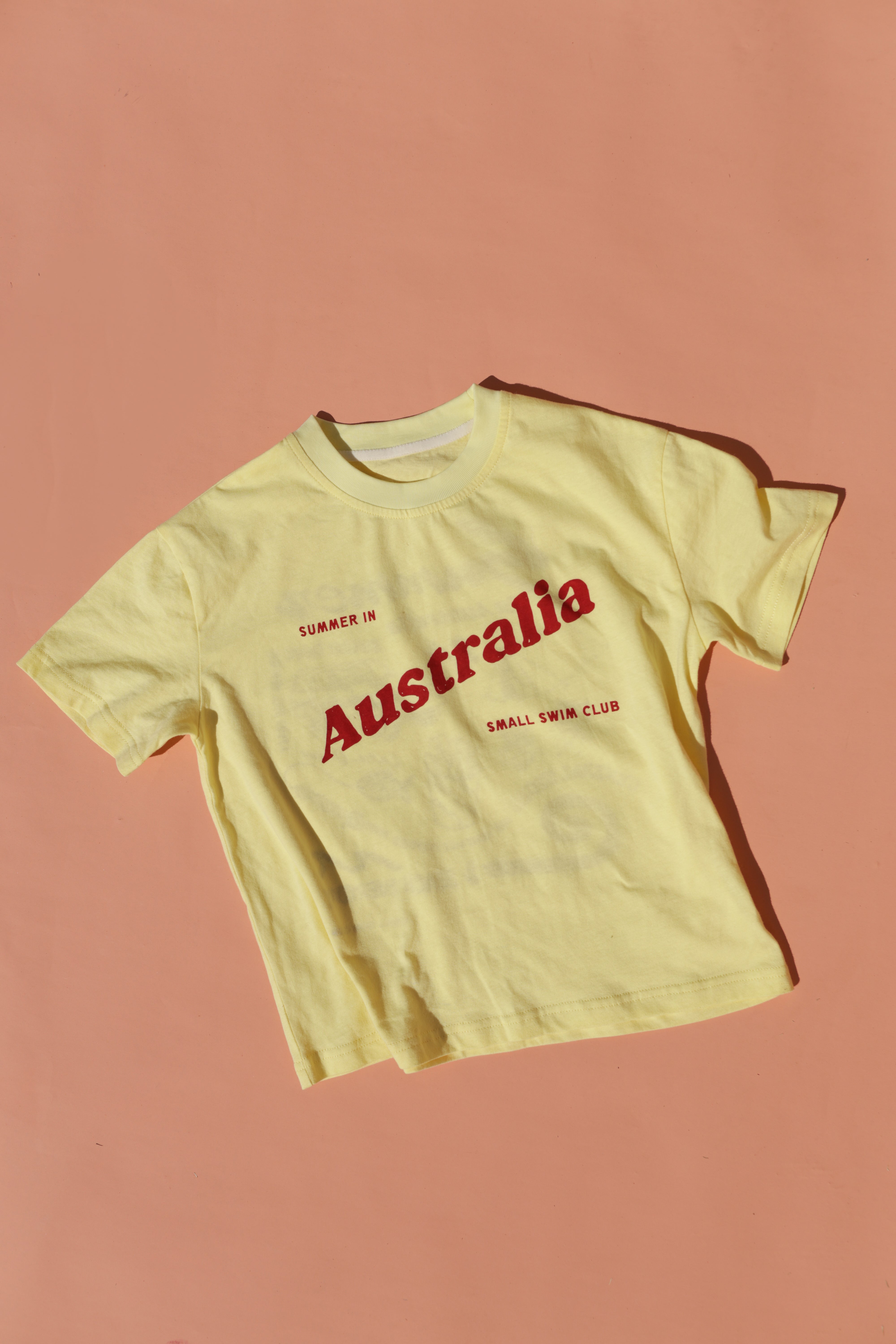 The Summer in Australia Tee Lemon by SMALL SWIM CLUB features maroon text on a yellow shirt, laid flat against a peach background. Made from 100% cotton, it's ideal for celebrating the Aussie summer with style and comfort.