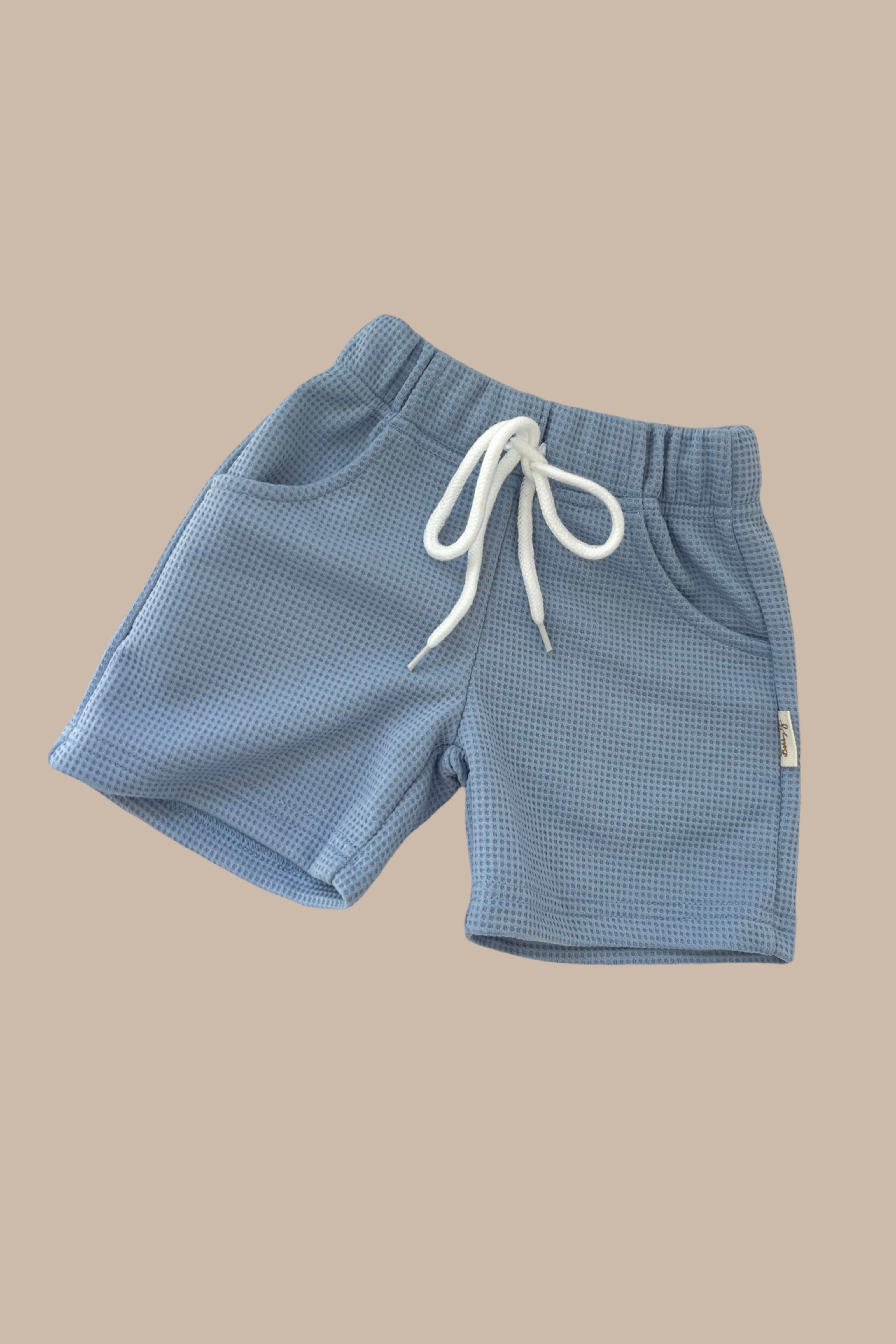 Introducing the Frankie Shorts Bleu by BOBBY G BABY WEAR: These light blue waffle knit jogger shorts offer a stylish and comfortable design. They feature an elastic waistband, white drawstring, and are crafted from luxe waffle fabric. The shorts include two front pockets and are showcased on a plain beige background.