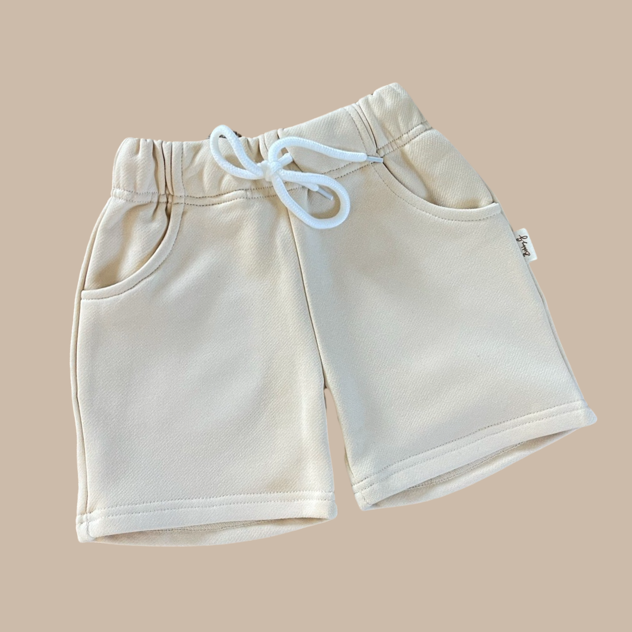 The Frankie Shorts Popcorn by BOBBY G BABY WEAR are beige toddler jogger shorts made from soft French terry fabric. They include a white drawstring and handy front pockets, all elegantly displayed on a light brown background.