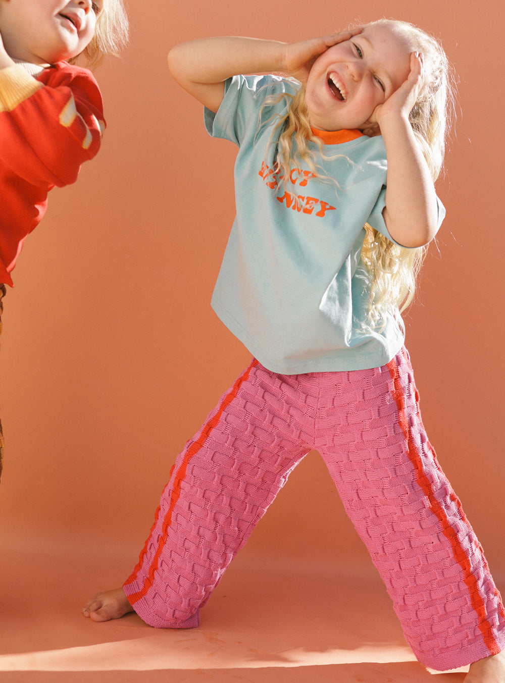 A young child with long hair is playfully posing, wearing a light blue T-shirt and the Twin Stripe Knit Pants in Fuschia/Red by SMALL SWIM CLUB, featuring a soft elastic waistband with orange stripes. Another child in a red top, reminiscent of lazy days at home, is partially visible, energetically jumping in the brightly lit room.