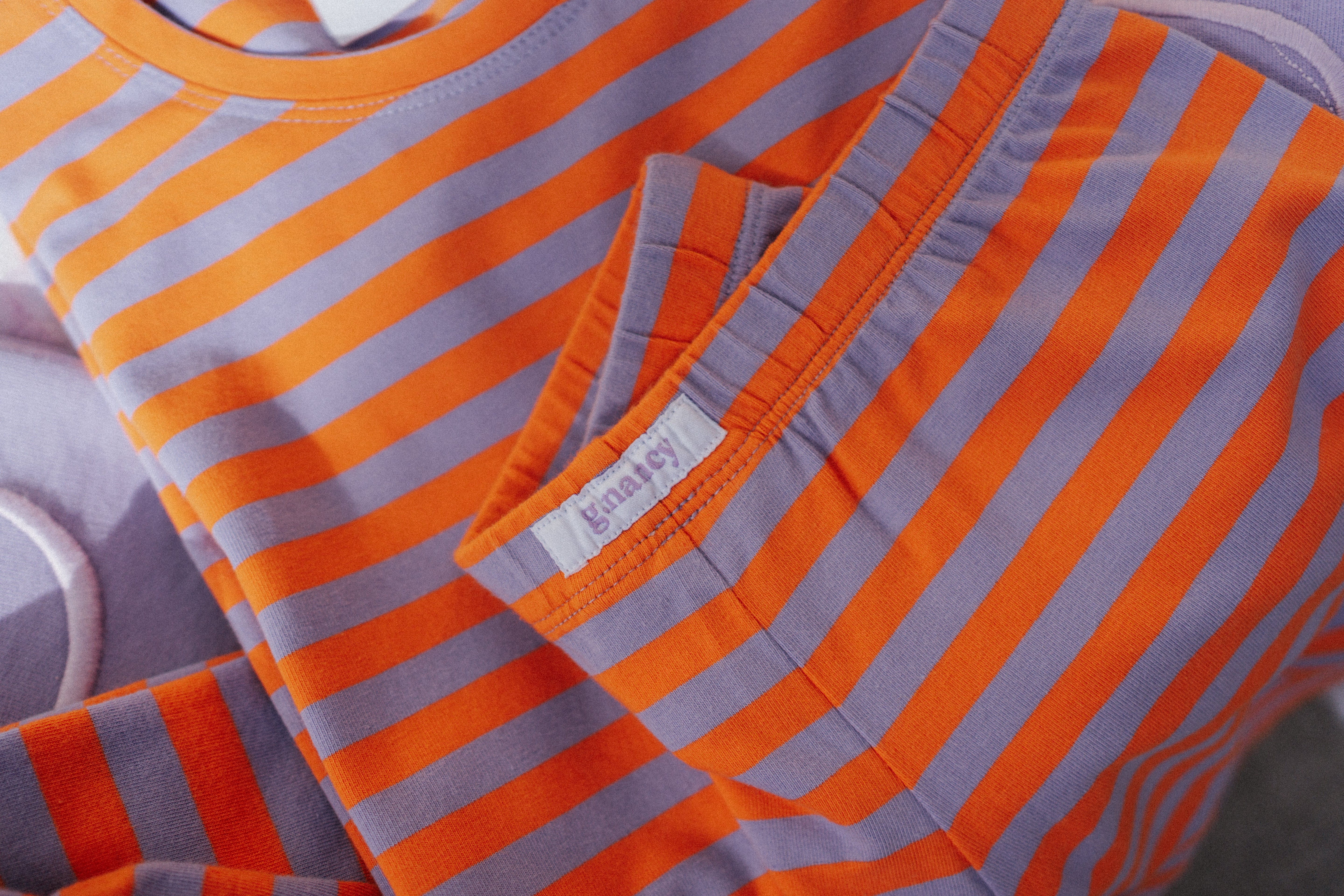 Close-up of the Stripe Shortie PJ Set Jacaranda/Peel by G.NANCY, featuring snug-fitting orange and purple stripes. A small white label is sewn onto the pocket. This limited edition piece is crafted from organic cotton jersey, showcasing a soft and neatly arranged fabric.