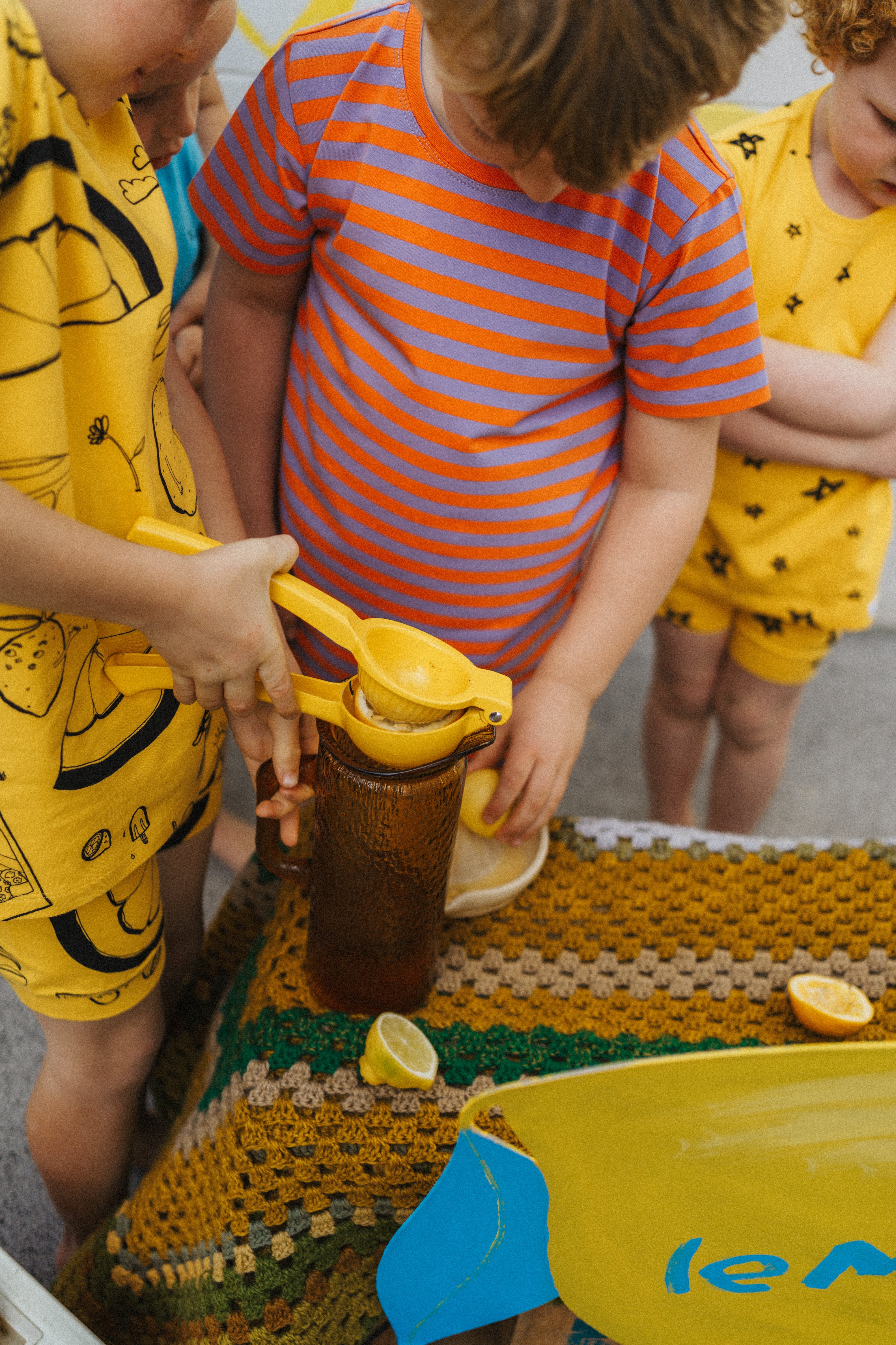 Children are using a yellow juicer to squeeze lemons into a glass jar while wearing the snug-fitting Stripe Shortie PJ Set in Jacaranda/Peel by G.NANCY. They gather around a table covered with a knit cloth, surrounded by scattered lemon slices. The scene captures the essence of summer's vibrant fun, like a limited edition moment.