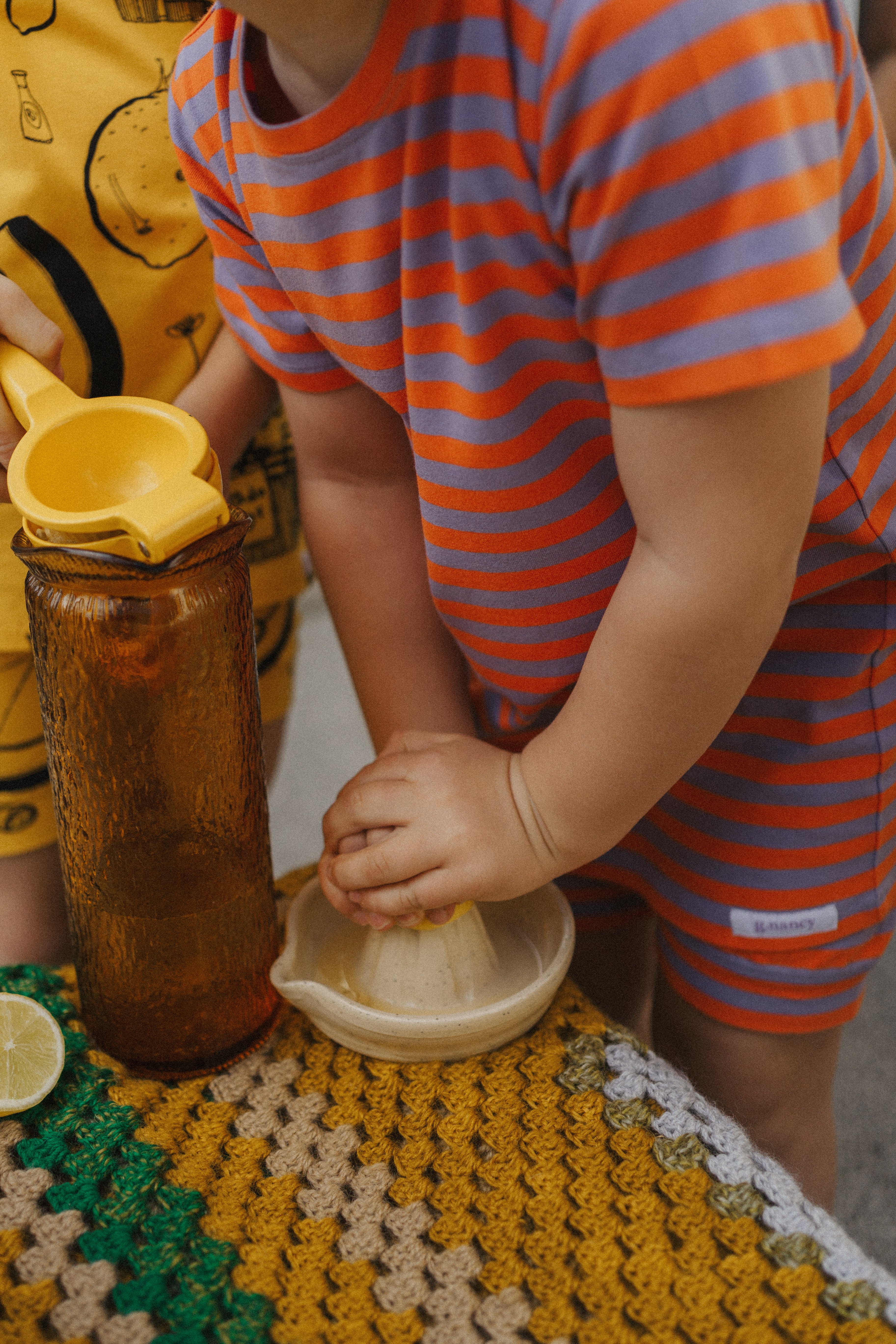 A child, comfortably dressed in the Stripe Shortie PJ Set Jacaranda/Peel by G.NANCY, uses a citrus juicer on a small table adorned with a knitted cloth. A yellow pitcher and a lemon half are nearby, completing this charming limited edition scene.