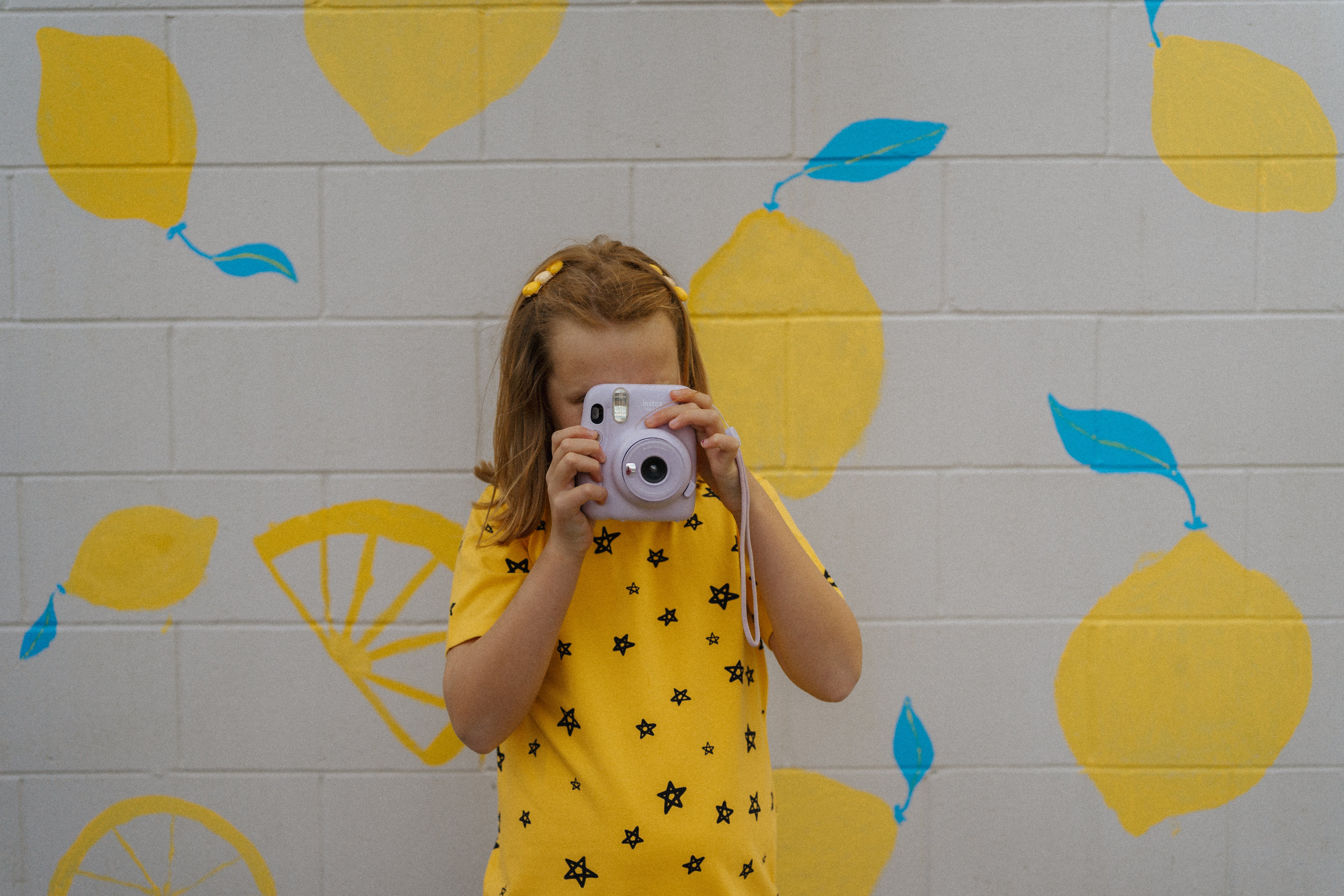 A young girl wearing the G.NANCY Star Shortie PJ Set Lemon, a cozy organic cotton sleepwear ensemble, holds a camera in front of her face. She stands against a gray brick wall adorned with yellow lemons and blue leaves, perfectly capturing the essence of comfort and charm.