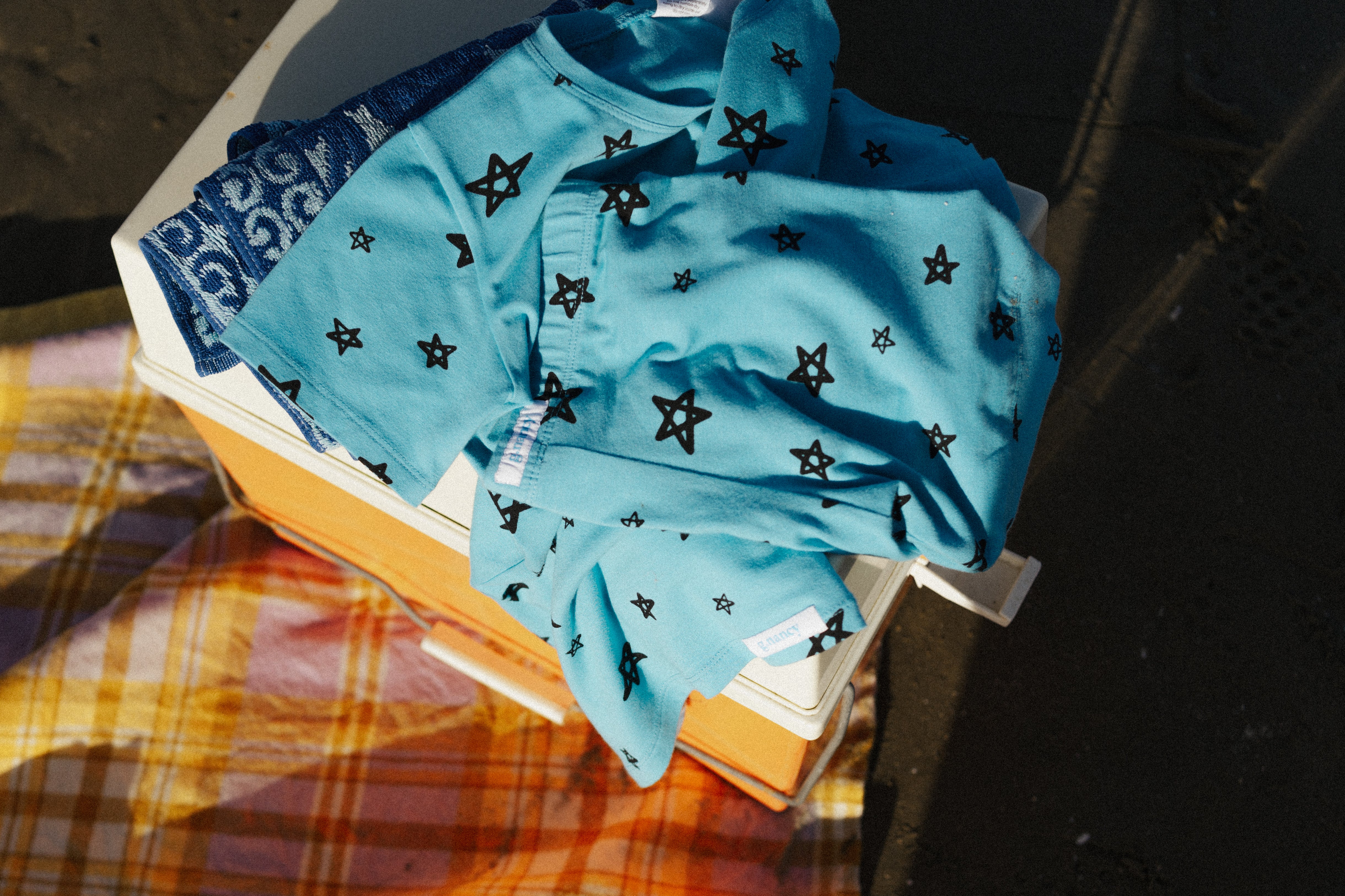 The Star Shortie PJ Set Breeze by G.NANCY, showcasing a hand-drawn star illustration, is draped over an orange cooler, enhanced by a blue patterned towel beneath it. A plaid blanket is laid out on the sand nearby, completing the cozy outdoor scene in the sunlight.