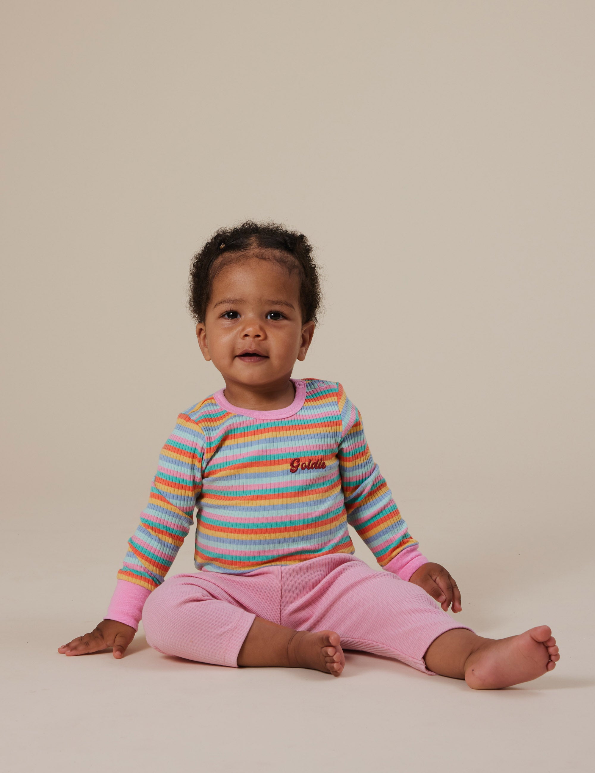 A baby sits calmly on the floor, wearing a colorful striped long-sleeve shirt and Goldie + Ace Bowie Rib Legging Sweet Pea. The pink pants have an elasticated waist and are made of a soft cotton/elastane blend for good stretch. The backdrop is a light, neutral color as the baby gazes at the camera.