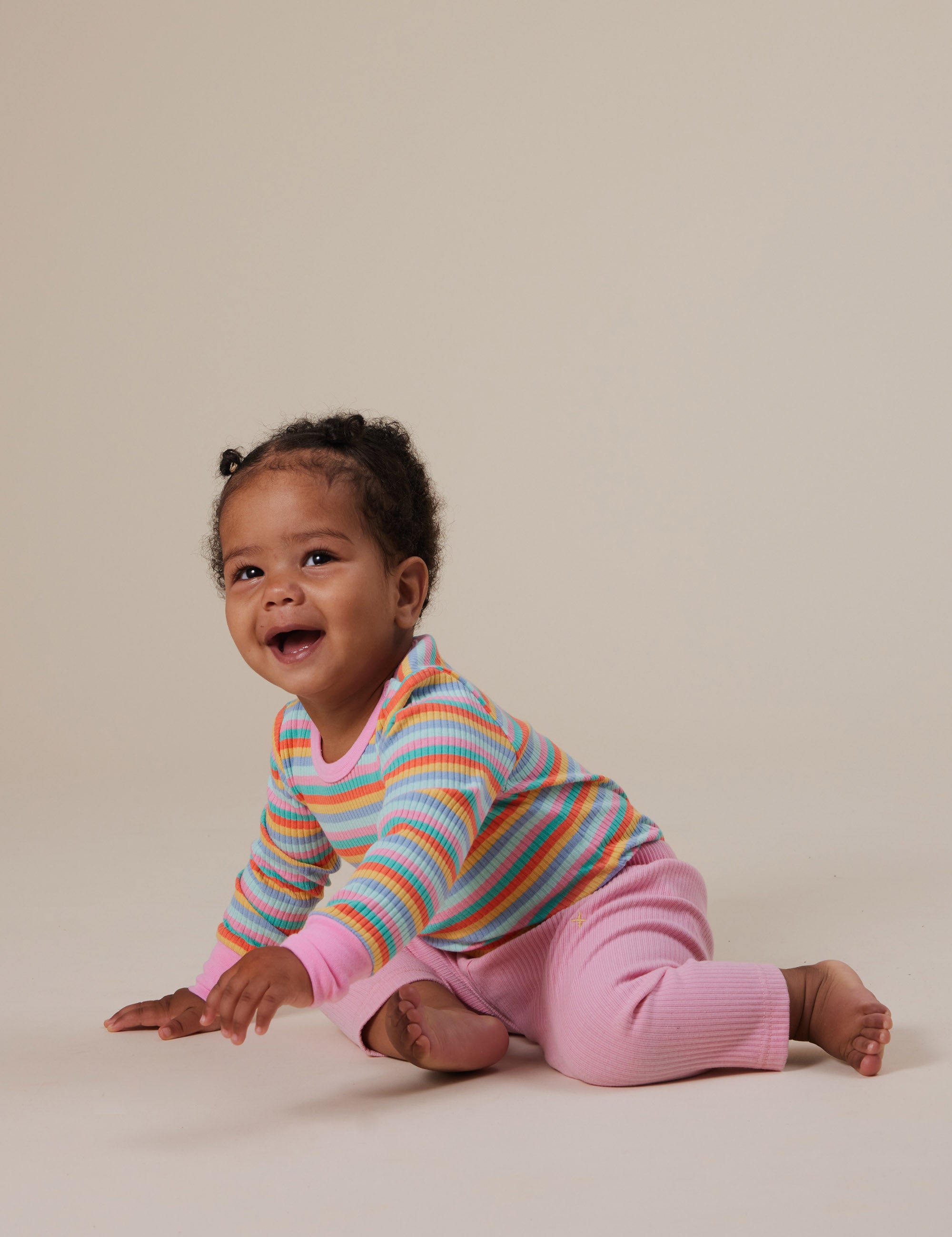 A baby in a colorful striped sweater and Goldie + Ace Bowie Rib Legging Sweet Pea by GOLDIE + ACE is sitting on the floor, smiling and looking up. The baby has short curly hair against a plain beige background.