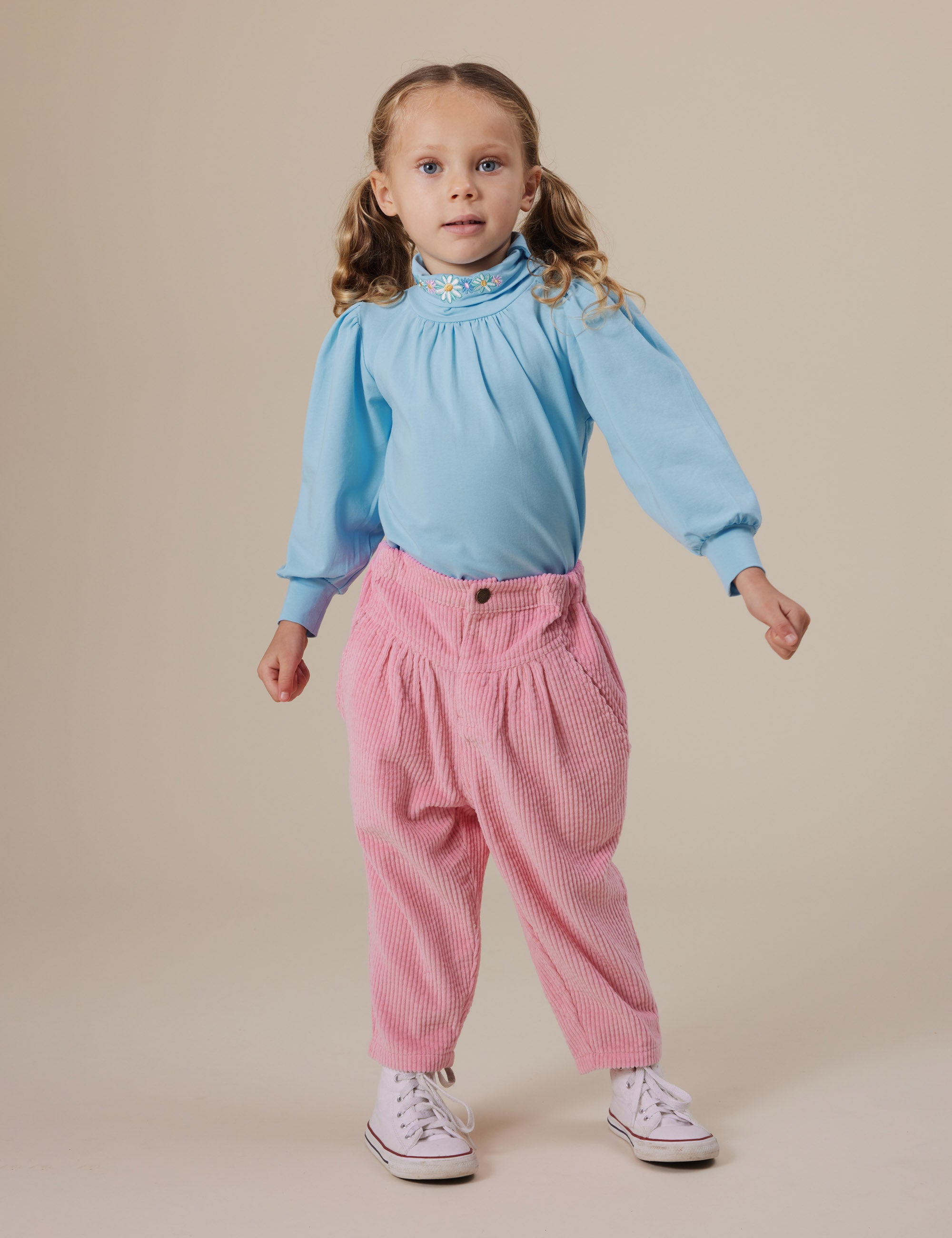 A young girl with light brown pigtails stands against a neutral backdrop wearing a light blue blouse, mid-rise pink Goldie + Ace ~ Nora Corduroy Pant from GOLDIE + ACE, and white sneakers. Her expression is neutral, with one arm slightly raised.