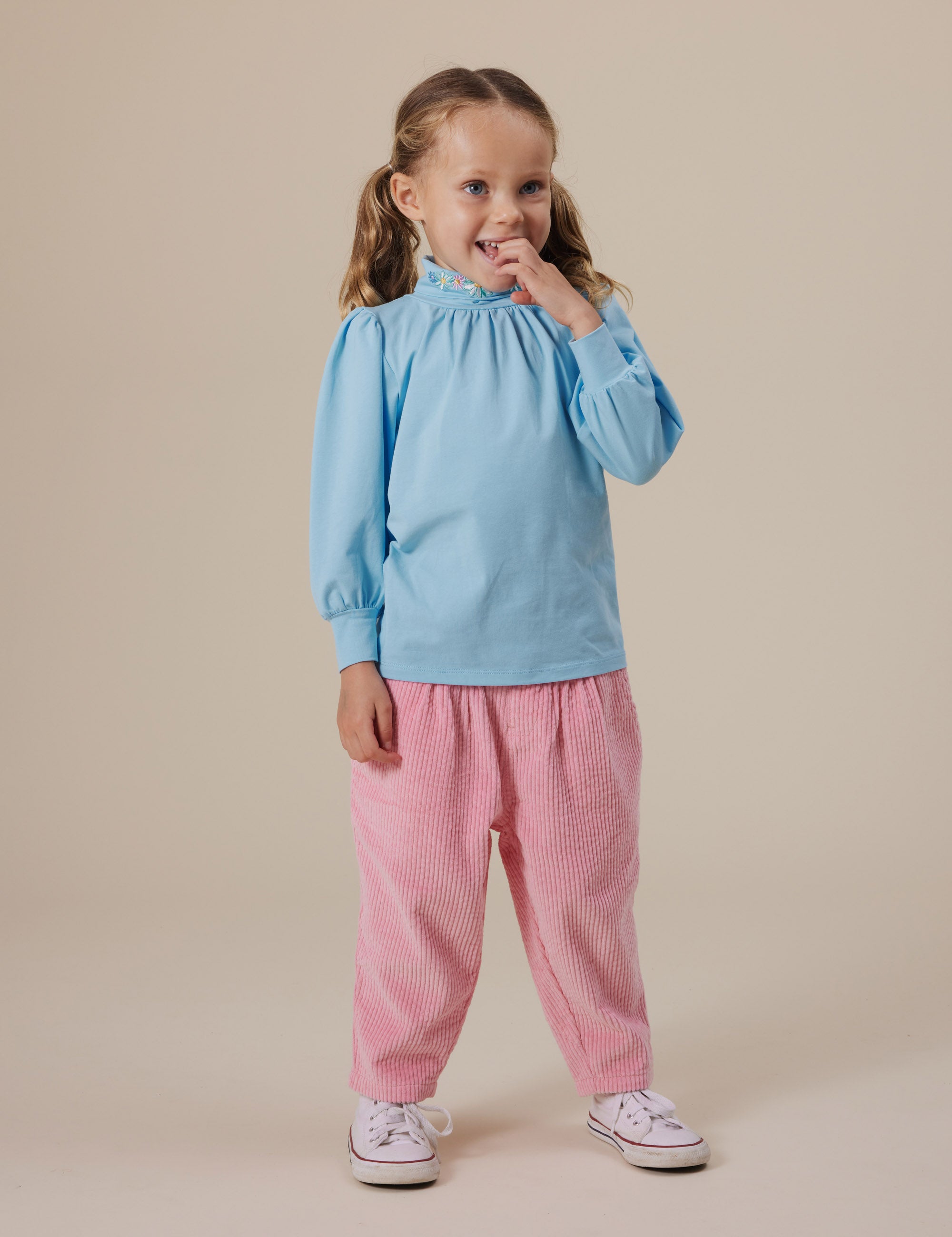 A young child stands against a neutral background wearing a light blue long-sleeve shirt and Goldie + Ace ~ Nora Corduroy Pant in pink with white sneakers. The retro style complements the child's brown hair in pigtails and their playful smile with a hand near their mouth.