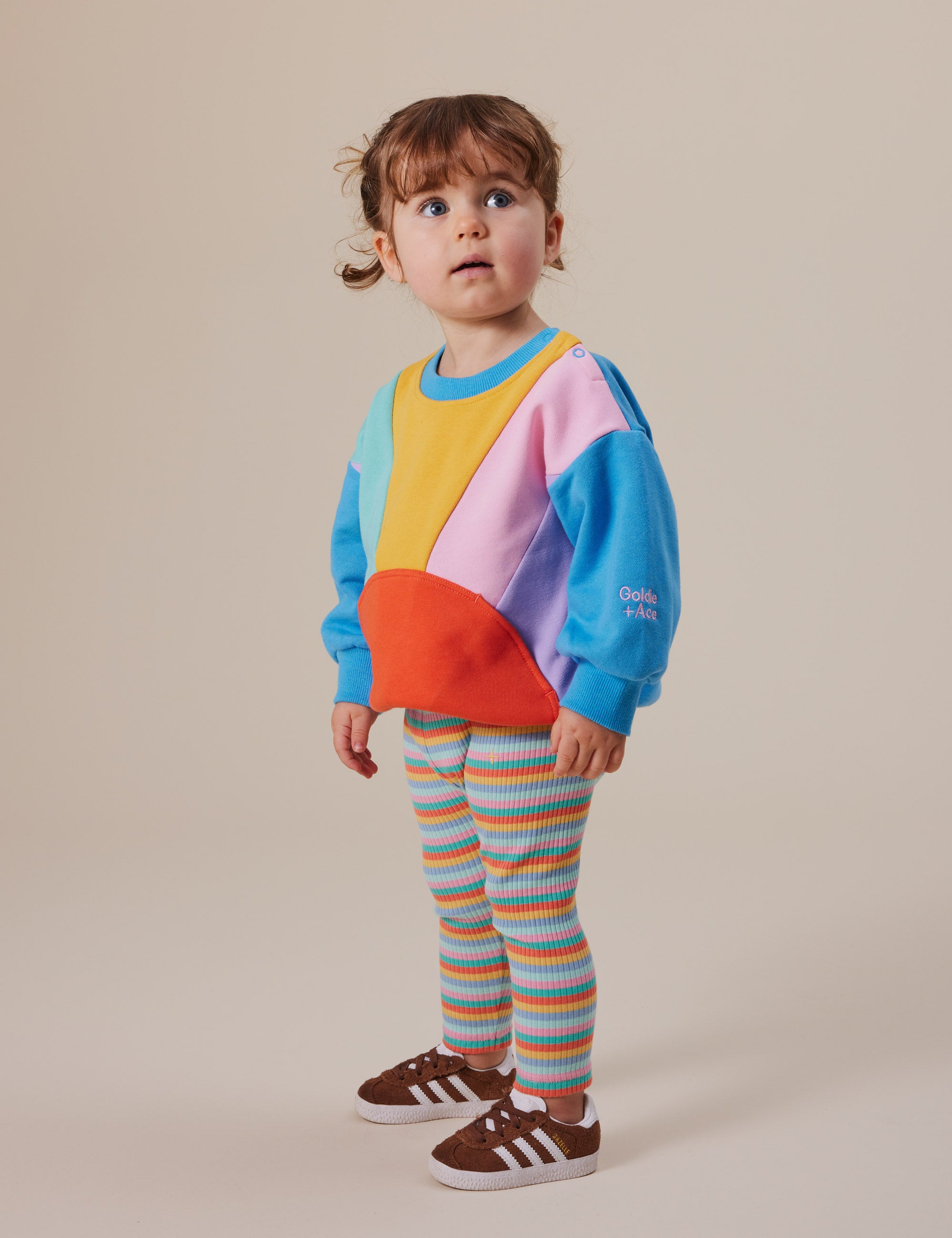 A young brown-haired child stretches comfortably in a colorful sweatshirt and Goldie + Ace Candy Stripe Rib Leggings with an elasticated waist. They wear brown sneakers against a plain beige background.