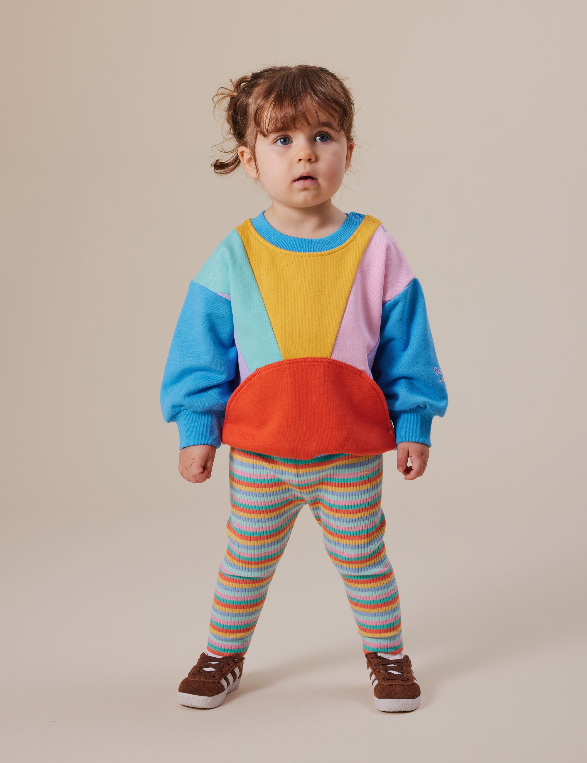 A toddler with brown hair in a ponytail stands against a plain background wearing colorful Goldie + Ace Candy Stripe Rib Leggings and a multi-colored cotton/elastane sweatshirt. The leggings feature an elasticated waist, and the child pairs the outfit with brown shoes.