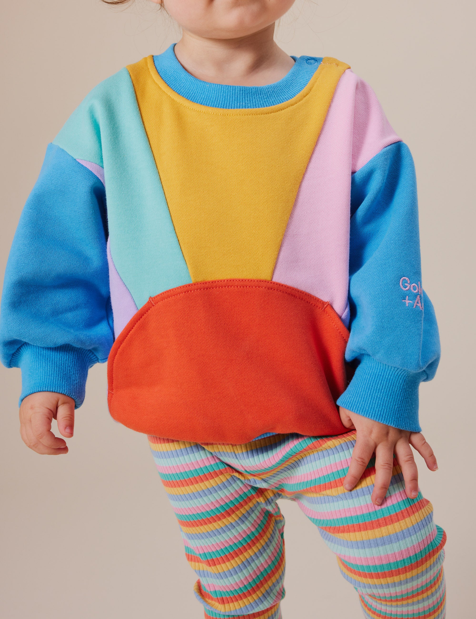 A young child models the Goldie + Ace sweatshirt featuring colorful yellow, blue, pink, and orange sections with "Going + AL" on the sleeve. Paired with GOLDIE + ACE Candy Stripe Rib Leggings boasting an elasticated waist for stretch and comfort, this outfit ensures ease for playful moves.