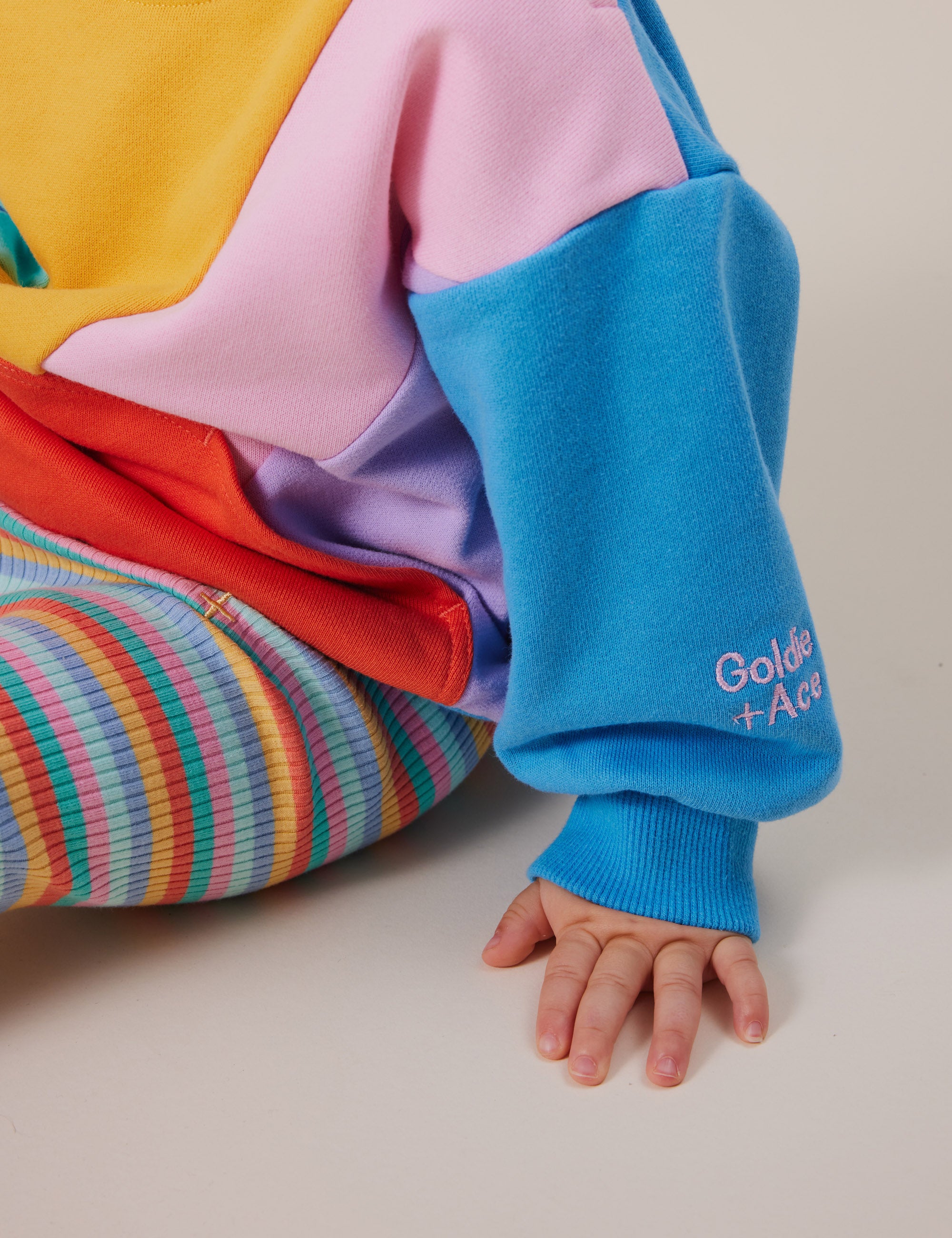 A child in a colorful outfit featuring blue sleeves with "Goldie+Ace" embroidery wears Goldie + Ace's Candy Stripe Rib Leggings. The pastel cotton/elastane ensemble offers good stretch, with striped pants and an elasticated waist as the child's hand gently touches the floor.