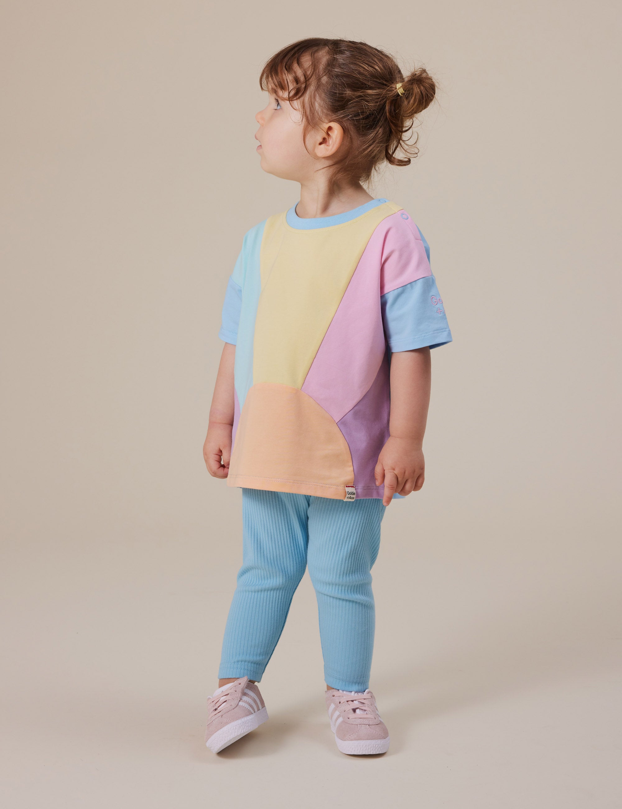 A young child with brown hair tied up stands sideways against a neutral backdrop, wearing a colorful pastel top made of cotton elastane for comfort and stretch, paired with Goldie + Ace's Bowie Rib Legging in Sky Blue featuring an elasticated waist and pink shoes.