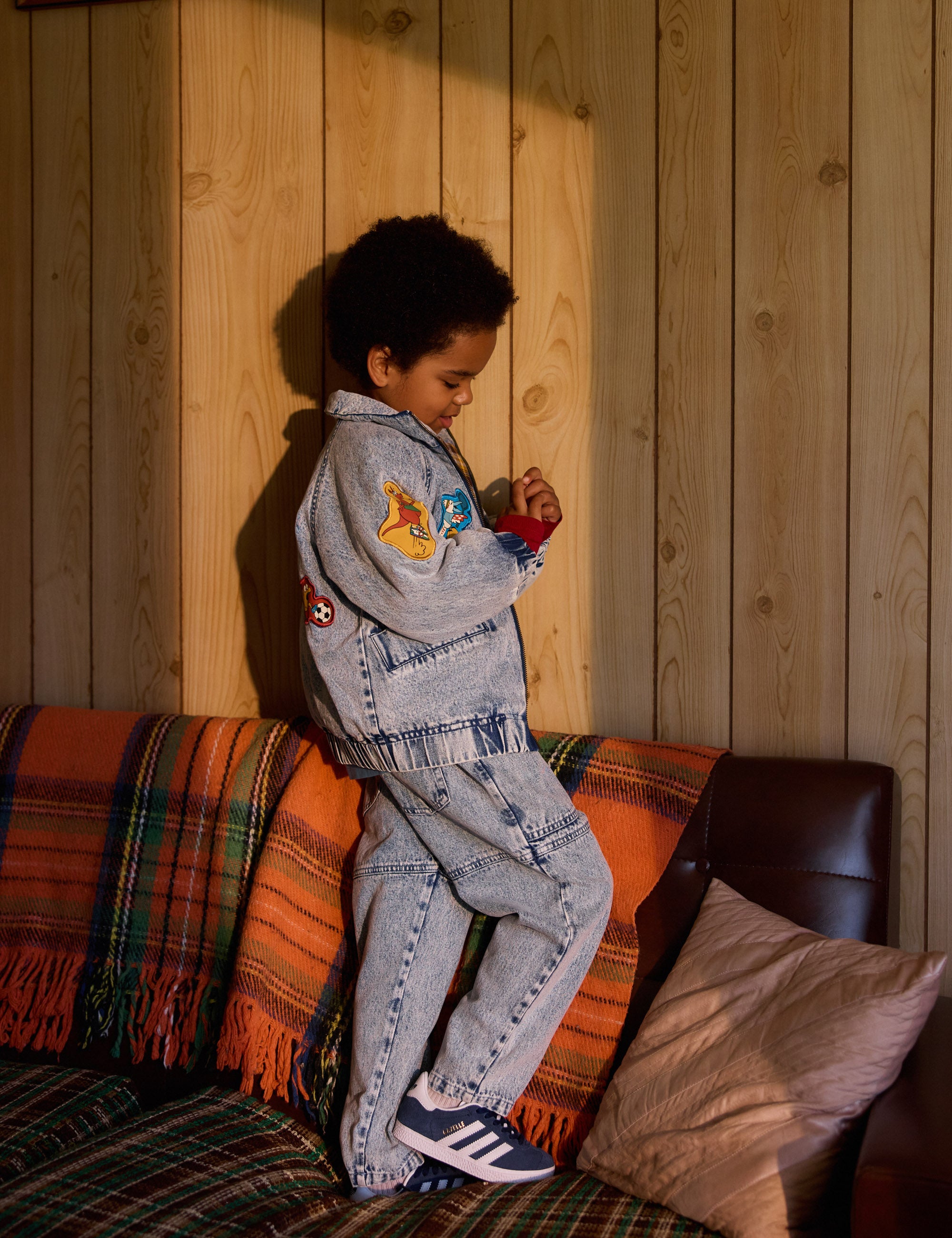 A child stands on a couch against a wooden wall, wearing Goldie + Ace's Drew Denim Pocket Pant in light denim with colorful patches. The couch, covered with a plaid blanket and beige cushion, complements the outfit as the child looks down while holding something.