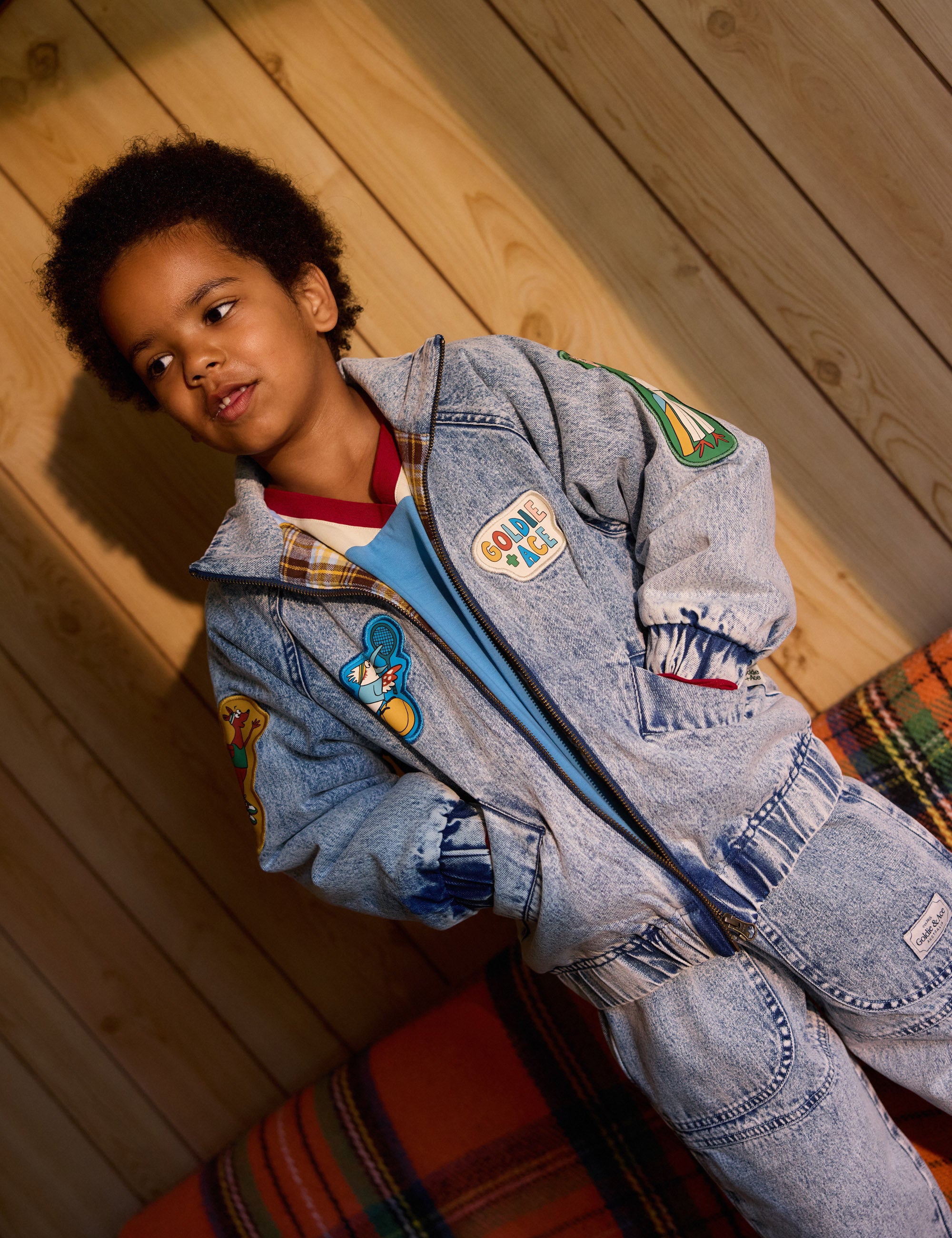 A young child stands against a wooden wall wearing the Goldie + Ace Drew Denim Pocket Pant in Light Denim. Wide legs with colorful cartoon patches complement their thoughtful expression, partially in shadow, as a striped blanket is visible on the nearby sofa.