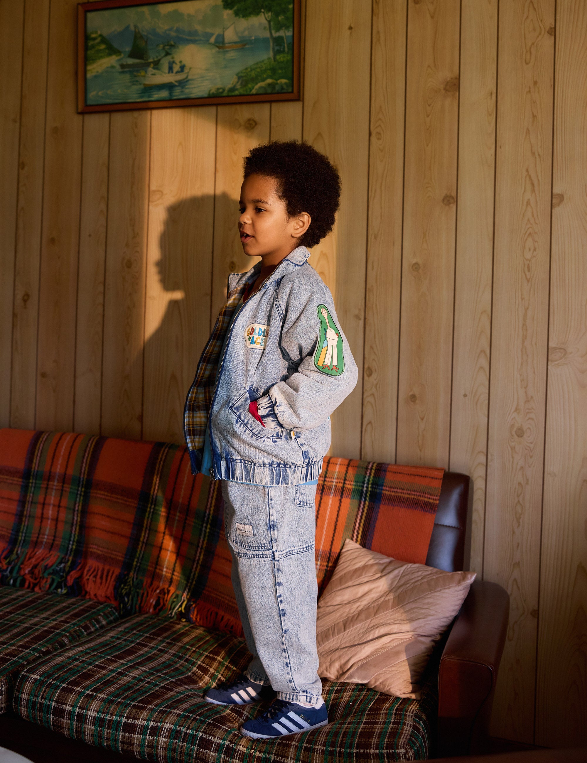 A child stands on a couch against wood-paneled walls, wearing a gray jacket and the stylish Goldie + Ace Drew Denim Pocket Pant in light denim with wide legs. The couch has colorful striped blankets and pillows, while a framed picture hangs above as the child gazes sideways, casting a shadow.