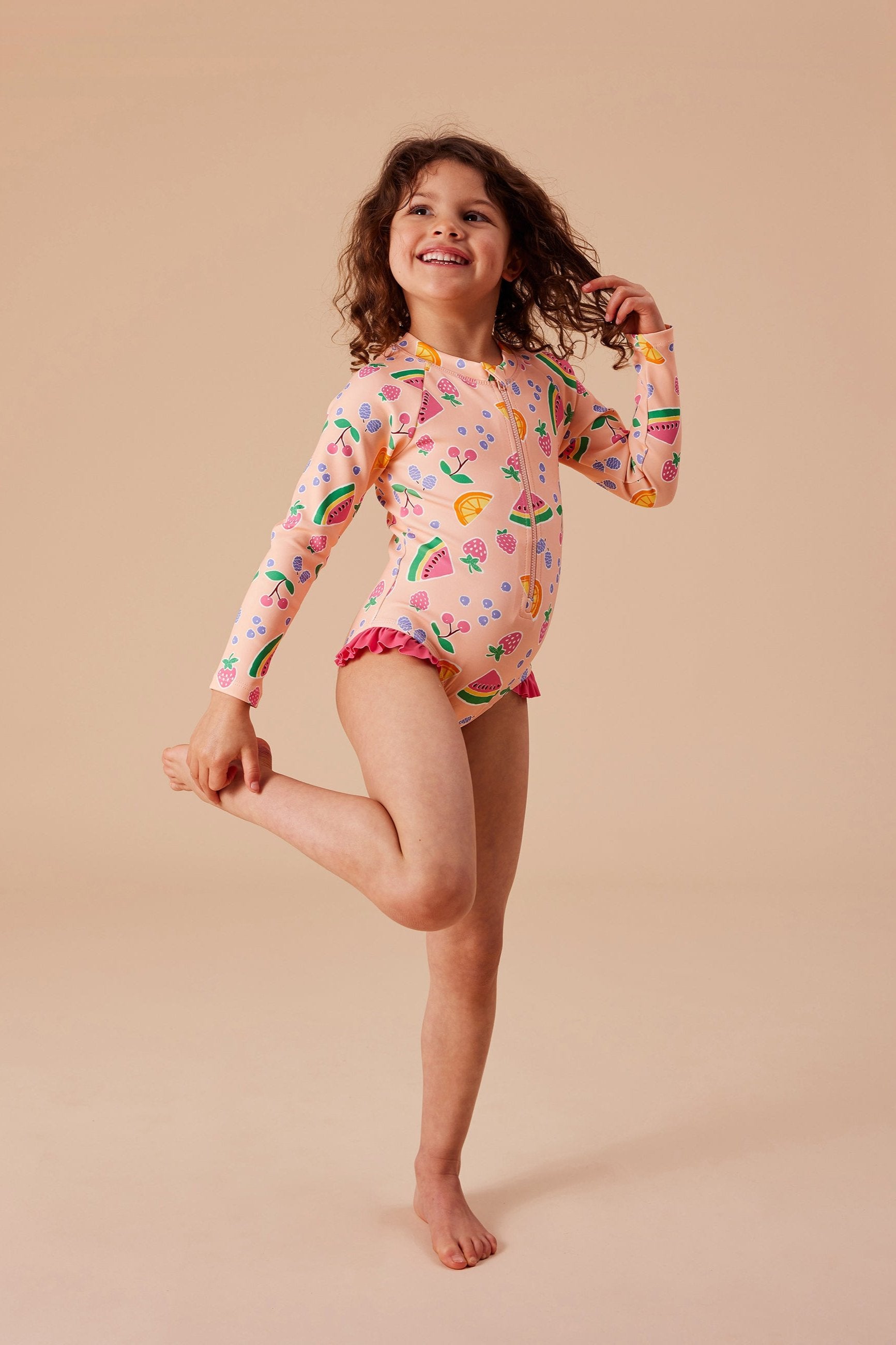 A young girl stands on one leg and smiles, dressed in the GOLDIE + ACE Fruit Salad Long Sleeve Swimsuit featuring vibrant, hand-drawn graphics of fruits and ice cream cones. Her other leg is bent at the knee, and her curly hair flows freely. The background is plain and beige. This adorable swimsuit offers SPF protection for carefree fun in the sun.