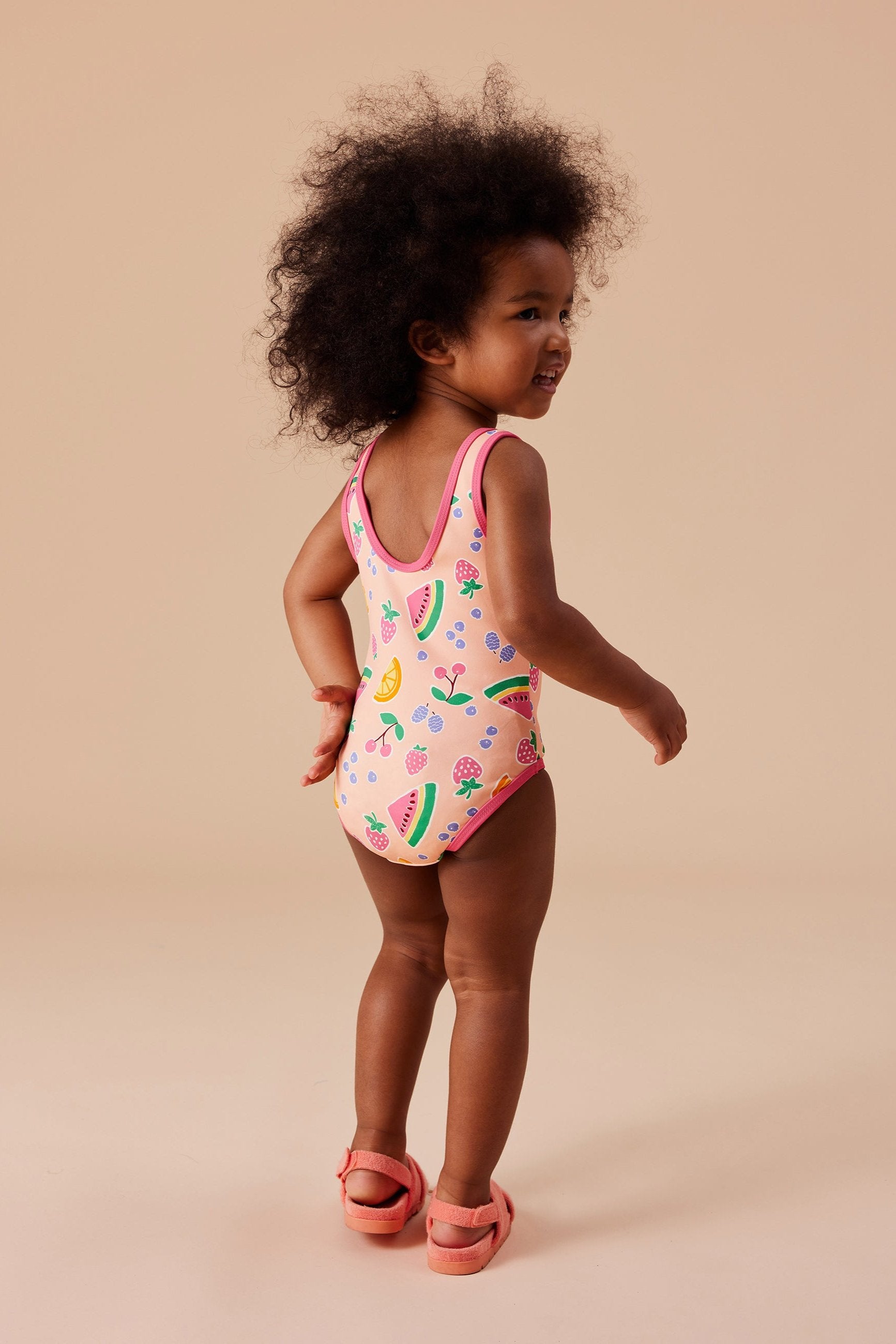 A toddler with curly hair stands on a beige background, smiling and looking to the side. The child is wearing GOLDIE + ACE’s Fruit Salad Scoop Back Bathers, a colorful swimsuit adorned with hand-drawn fruit patterns made from nylon/spandex material, and pink sandals.