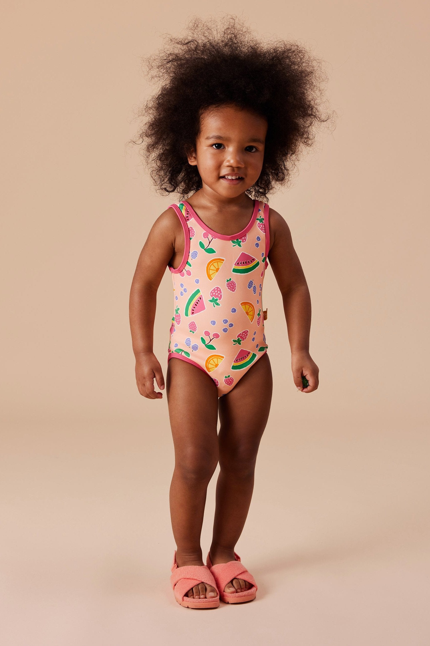 A young child with curly hair stands smiling in GOLDIE + ACE's Fruit Salad Scoop Back Bathers, showcasing colorful fruit prints and hand-drawn graphics. The child completes the look with matching pink sandals, all true to size, set against a simple beige background.