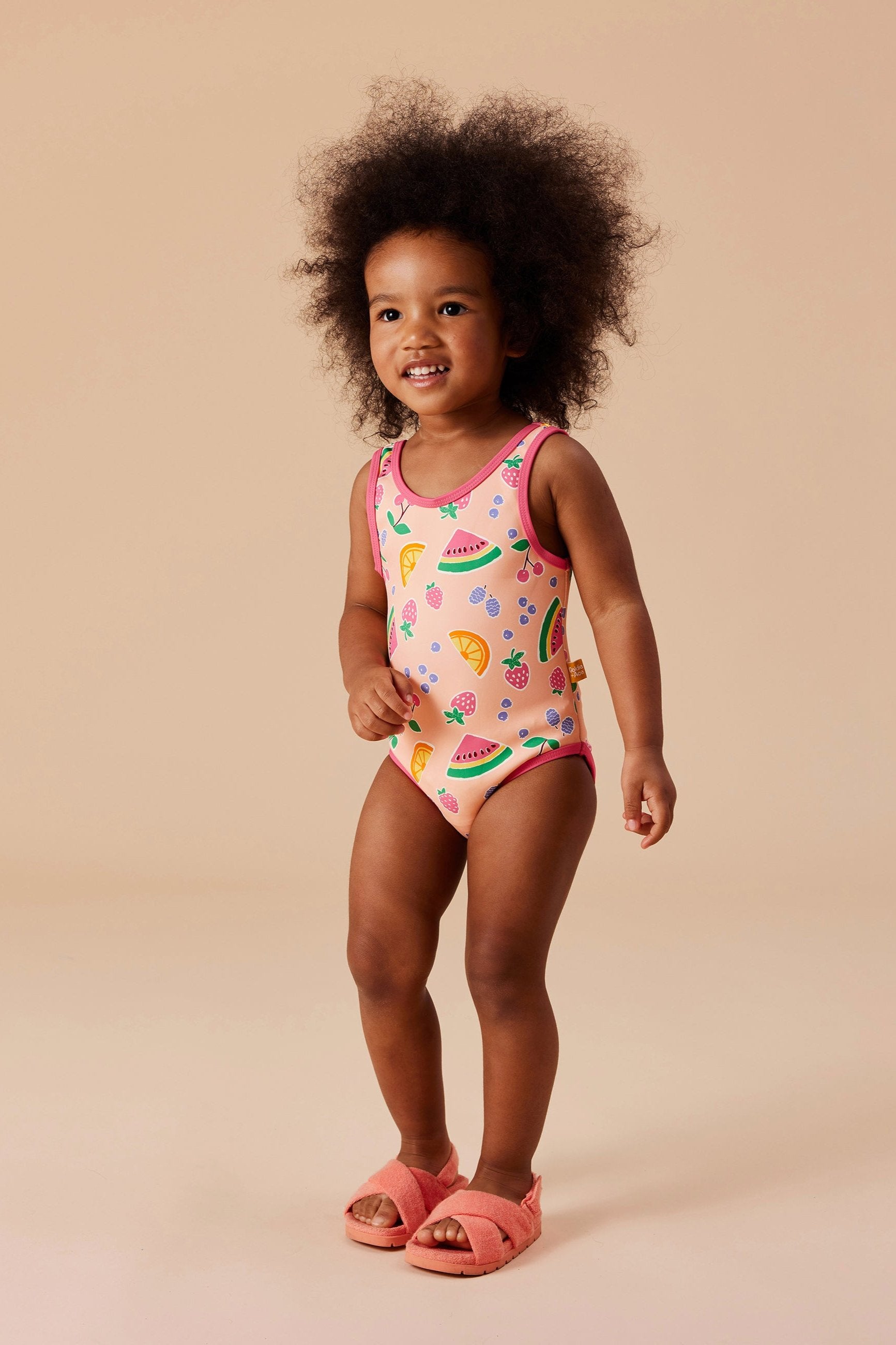 A toddler with curly hair stands and smiles, wearing the vibrant "Fruit Salad Scoop Back Bathers" by GOLDIE + ACE, made of nylon/spandex and adorned with hand-drawn graphics of watermelons, lemons, and strawberries. The child also wears pink sandals against a neutral beige backdrop.