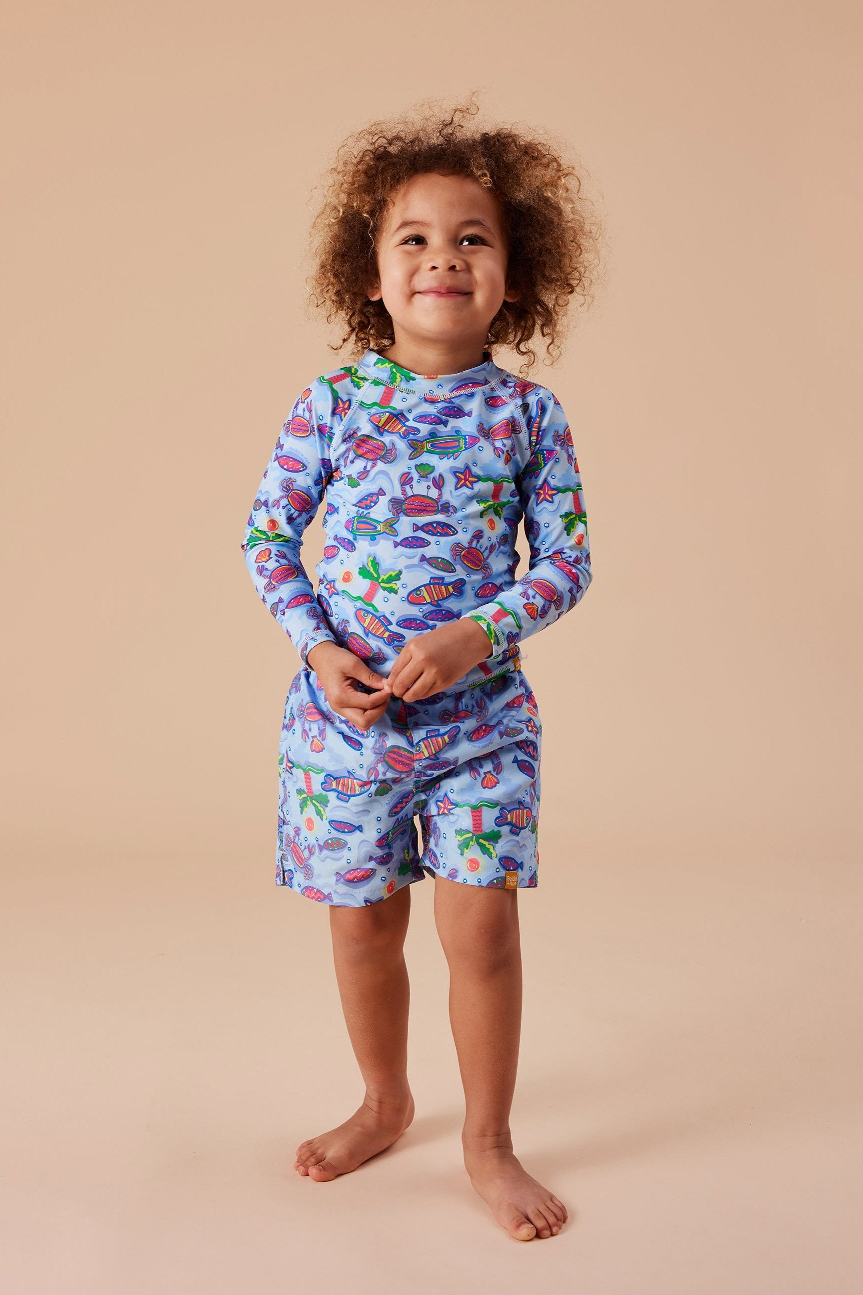 A young child stands against a plain beige background, smiling. They have curly hair and are wearing GOLDIE + ACE's Daydream Island Board Shorts, a long-sleeve blue/purple multi two-piece outfit with colorful cartoon animals and sea creatures. The shorts feature a functional drawstring, completing the matching set made from soft microfibre twill.