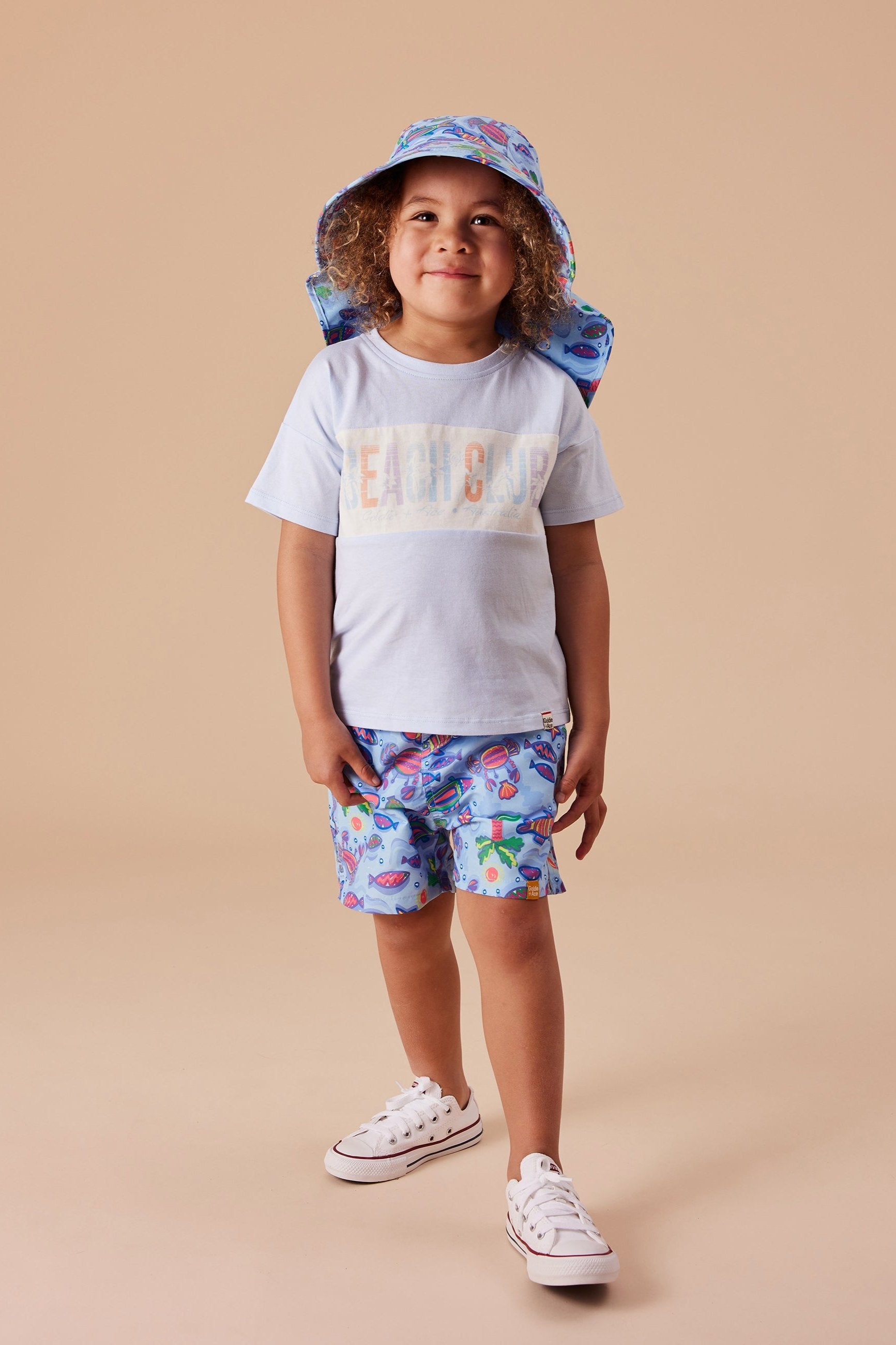 A child with curly hair stands smiling, wearing a light blue T-shirt with "BEACH CLUB" written on the front, Daydream Island Board Shorts by GOLDIE + ACE made of microfibre twill with vibrant fish patterns, a matching fish-patterned bucket hat, and white sneakers. The background is a plain beige color.