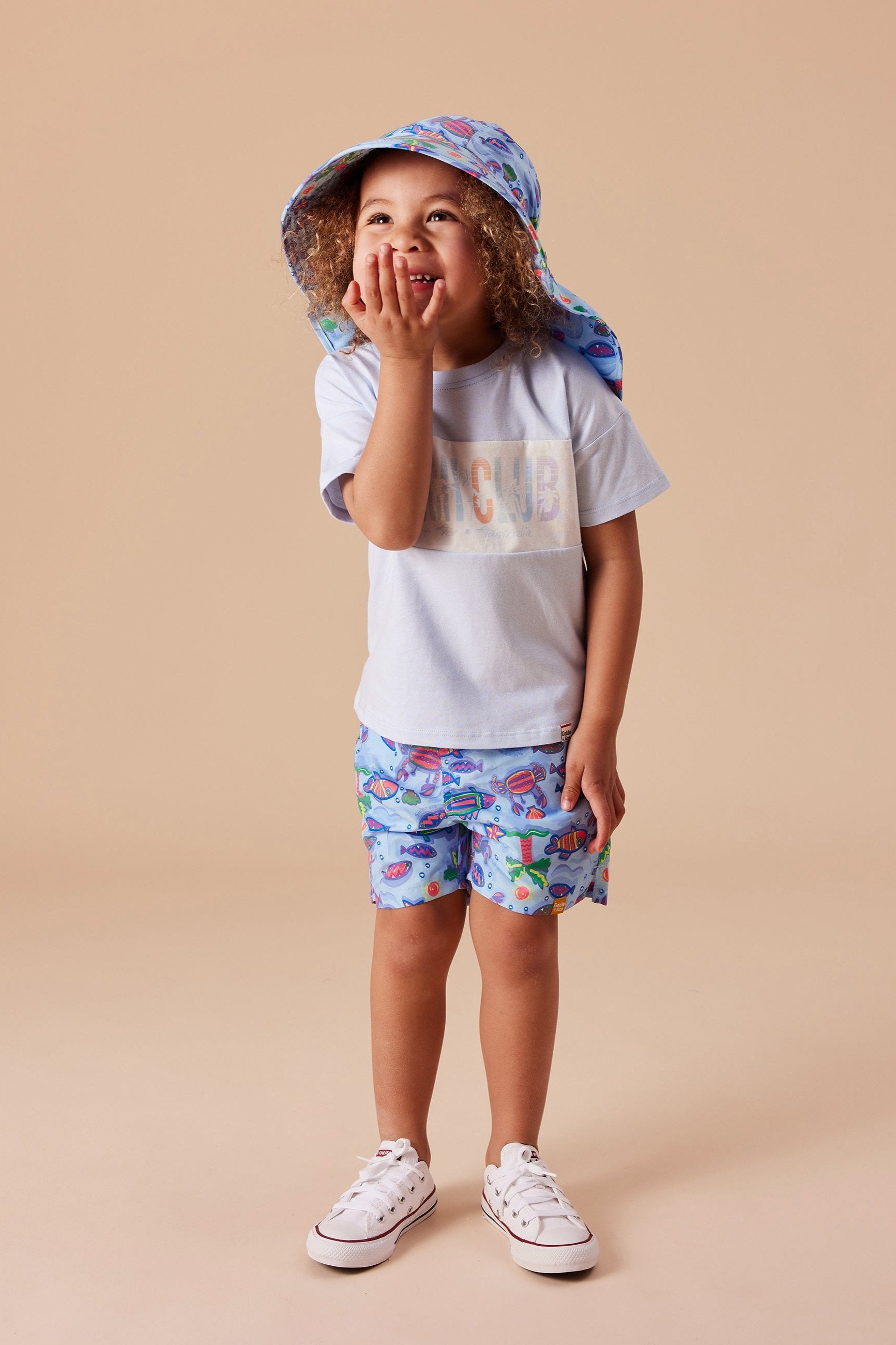 A young child sporting a light blue hat, GOLDIE + ACE Daydream Island Board Shorts in Blue/Purple Multi with colorful patterns and a functional drawstring, and a light grey T-shirt stands against a beige background. The child has a joyful expression, with one hand covering their mouth and the other resting on their shorts.