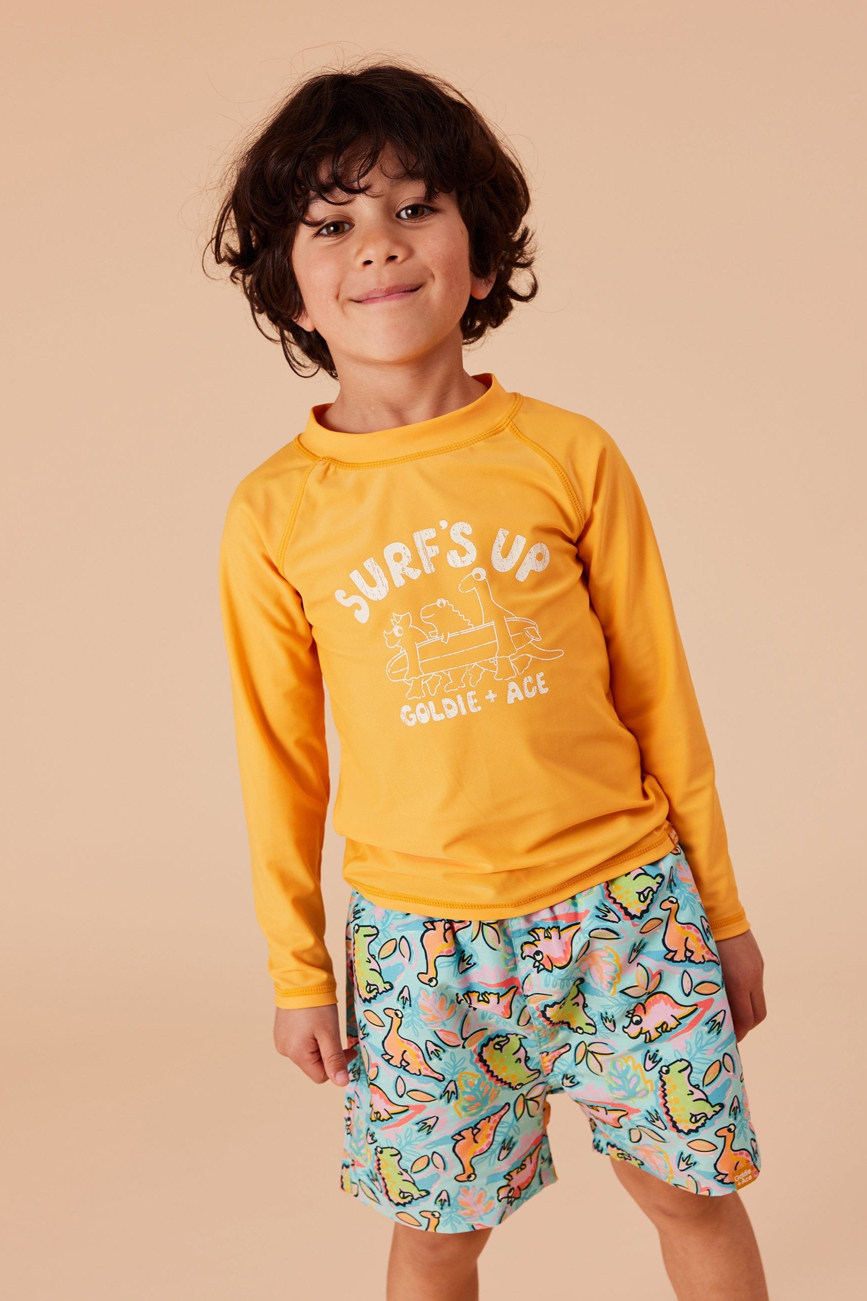 A young boy with curly hair is smiling while wearing a bright yellow long-sleeve shirt that says "Surf's Up" and GOLDIE + ACE's Dino Jungle Board Shorts in Aqua Green Multi, featuring a playful dinosaur pattern. He stands against a plain, tan background, showcasing his fun and true-to-size style.