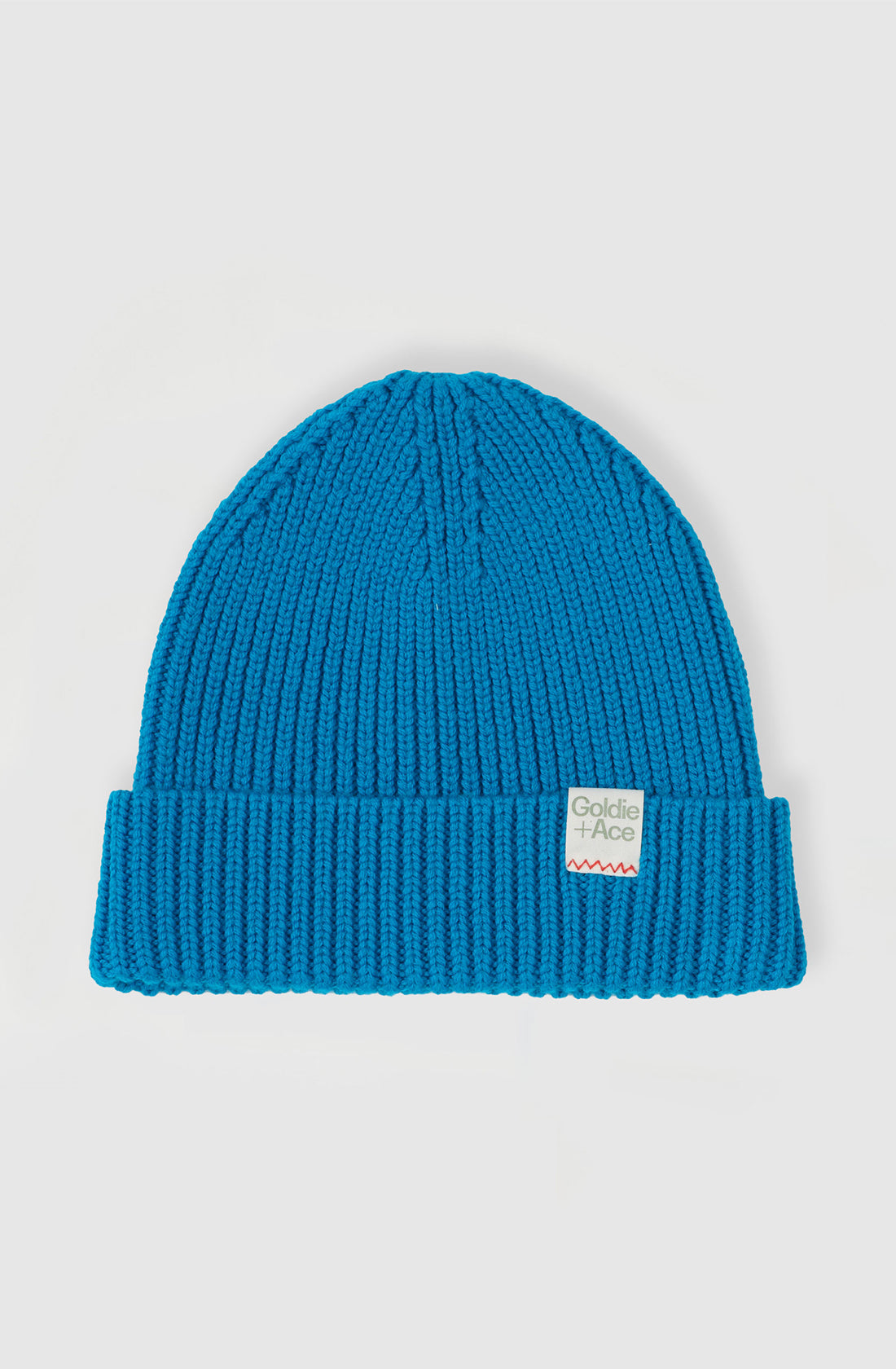 The Wool Beanie Lake from GOLDIE + ACE is a blue knit rib beanie featuring a folded cuff adorned with a small rectangular patch displaying "Goldie + Ace" and a red zigzag line below. Made from pure wool, this beanie offers a generous fit and is showcased on a plain white background.