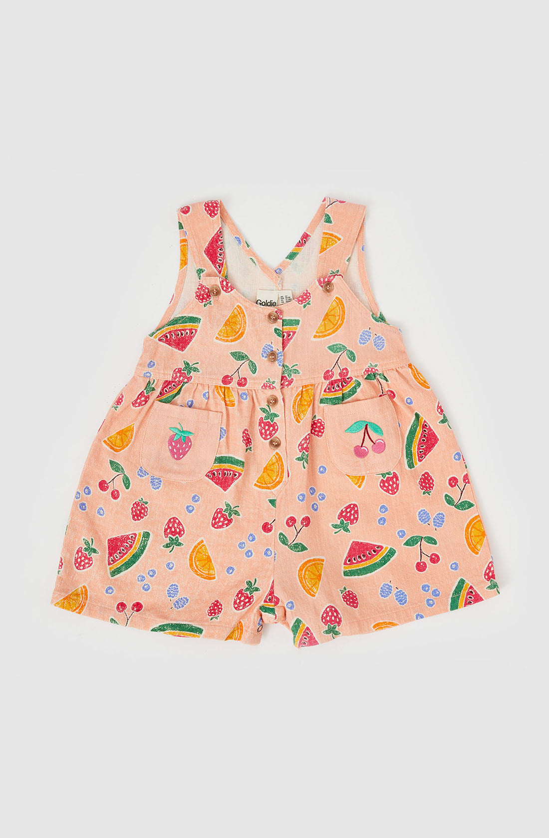 The Sara Linen Shortalls from GOLDIE + ACE is a light pink romper for toddlers, showcasing a hand-drawn print of colorful fruits such as watermelons, lemons, strawberries, and cherries. The shortalls feature adjustable button straps and a front pocket with a gathered waist for an adorable and playful appearance.