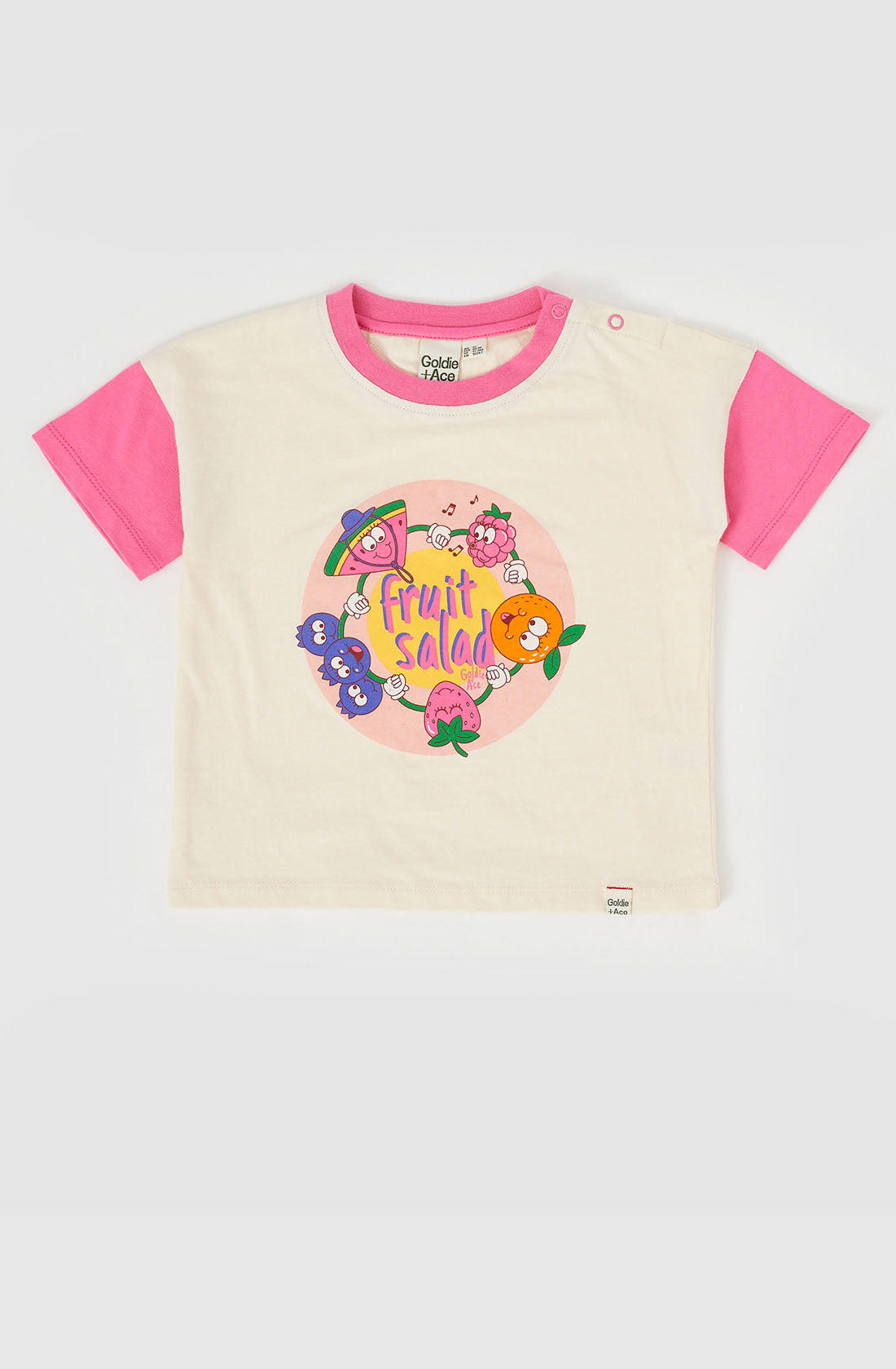 A short-sleeve, white children's t-shirt with pink sleeves from GOLDIE + ACE. The front features a colorful hand-drawn print of various cartoon fruits and text that reads "Fruit Salad." Made from super soft stretch cotton jersey, the shirt has a small brand tag at the bottom hem. This item is named the Fruit Salad Friends T-shirt.