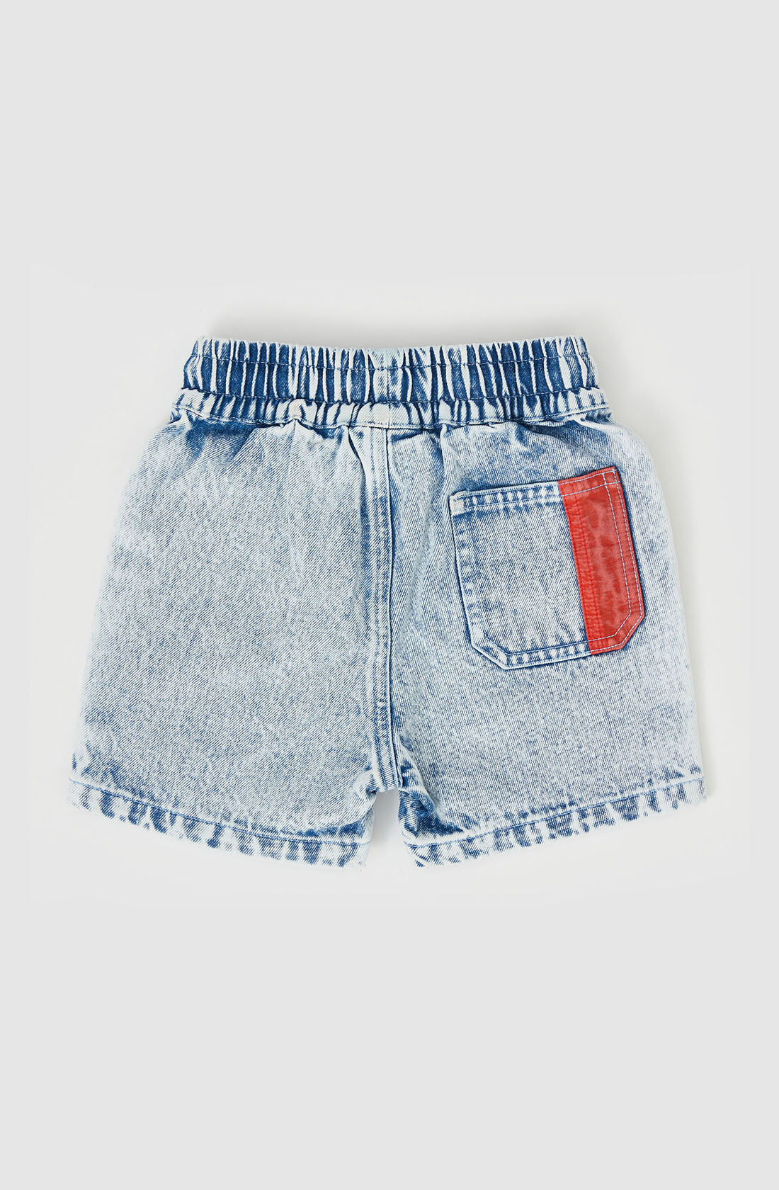 The Noah Colour Pop Denim Shorts by GOLDIE + ACE are shown from the back. These light denim shorts feature an elastic waistband with dark blue stitching, a functional drawcord, and one back pocket on the right side adorned with a red rectangular patch. The shorts have a slightly faded look, adding to their casual style.