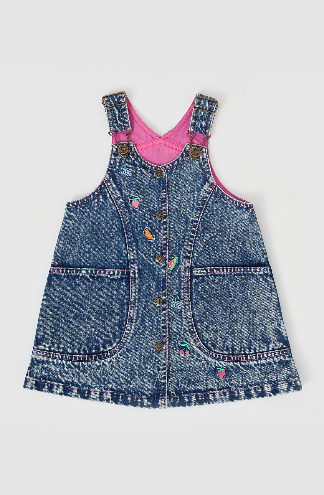 The Penelope Fruity Denim Pinafore Dress by GOLDIE + ACE is a sleeveless dark denim dress with two large front pockets and a button-down front. It features hand-drawn embroidery of small fruits and hearts. The inside lining is bright pink, while the adjustable brass straps add a touch of elegance.