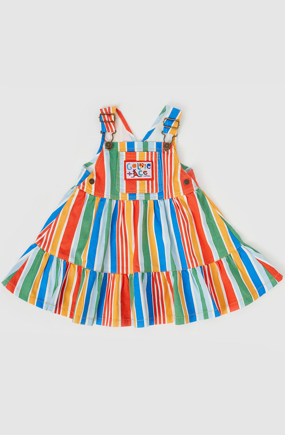 Introducing the Tully Tiered Pinafore Dress by GOLDIE + ACE: a bright, striped dress for kids featuring vivid vertical stripes in red, green, blue, orange, and yellow. This sleeveless dress showcases a flared skirt section and adjustable straps with brass snap buttons. A small front pocket is adorned with the playful words "Cool Kids.