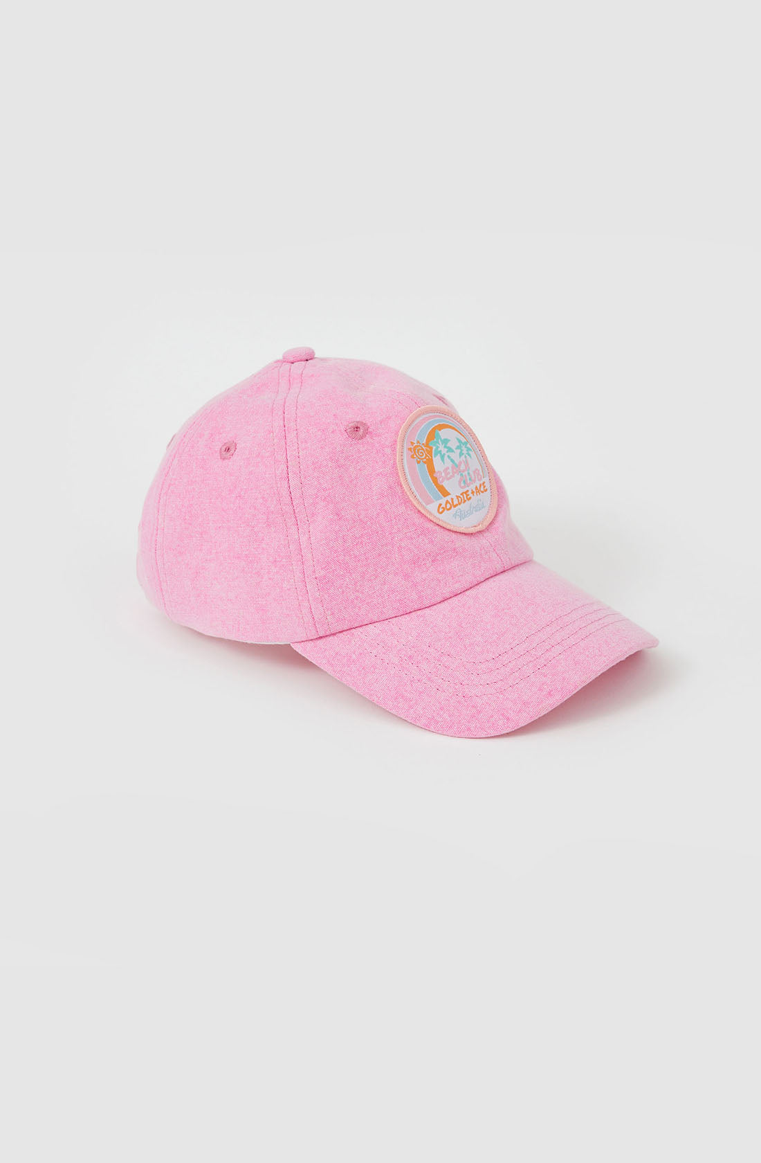 The Tate Beach Club Denim Cap by GOLDIE + ACE is a light pink cap crafted from pink washed denim. It features a colorful round embroidered patch on the front, displaying an illustration of three cacti with the word "California" beneath it. The cap includes a curved brim, button top, and an adjustable strap to ensure a perfect fit. The background is plain white.