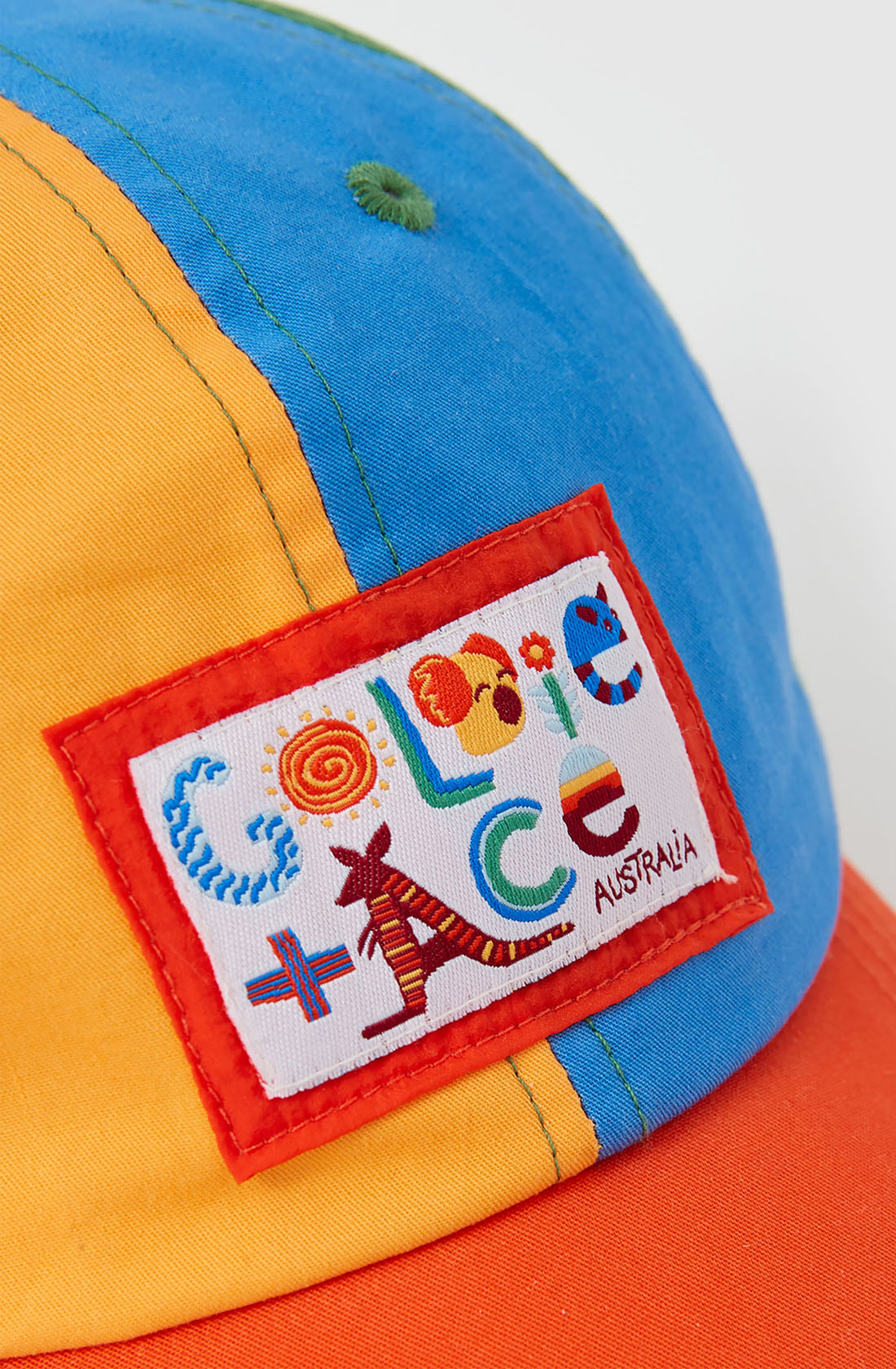 The Tate Land Down Under Denim Cap by GOLDIE + ACE is showcased with vibrant panels of blue, yellow, and orange fabric. It boasts a lively embroidered patch displaying the text "COLLAGE AUSTRALIA" along with a design featuring a kangaroo, lollipop, and various playful shapes. The cap includes an adjustable strap to ensure a perfect fit for any wearer.