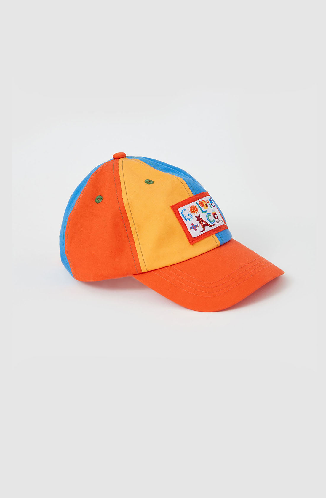 The Tate Land Down Under Denim Cap by GOLDIE + ACE is a colorful hat featuring an orange visor with blue, yellow, and orange panels. It sports a front patch adorned with playful, multicolored designs, showcasing various small shapes and symbols. Additionally, the cap boasts an adjustable strap for a true-to-size fit.