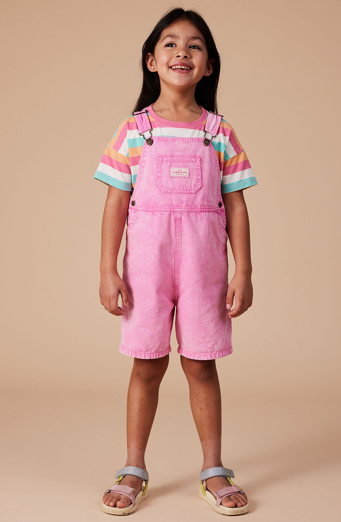 A young child with long dark hair smiles while wearing the Burton Vintage Washed Denim Overalls in light pink from GOLDIE + ACE. The overalls feature adjustable shoulder straps and are styled over a striped T-shirt in pastel colors. They are also wearing colorful sandals and standing against a beige background.