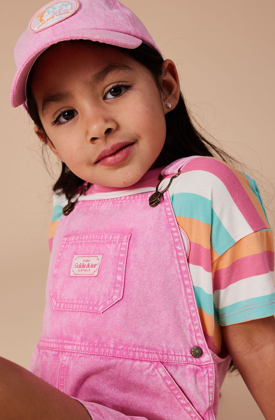 A young girl is wearing GOLDIE + ACE's Burton Vintage Washed Denim Overalls with adjustable shoulder straps, a colorful striped T-shirt, and a pink cap. She is looking at the camera with a neutral expression. The background is a plain beige color.