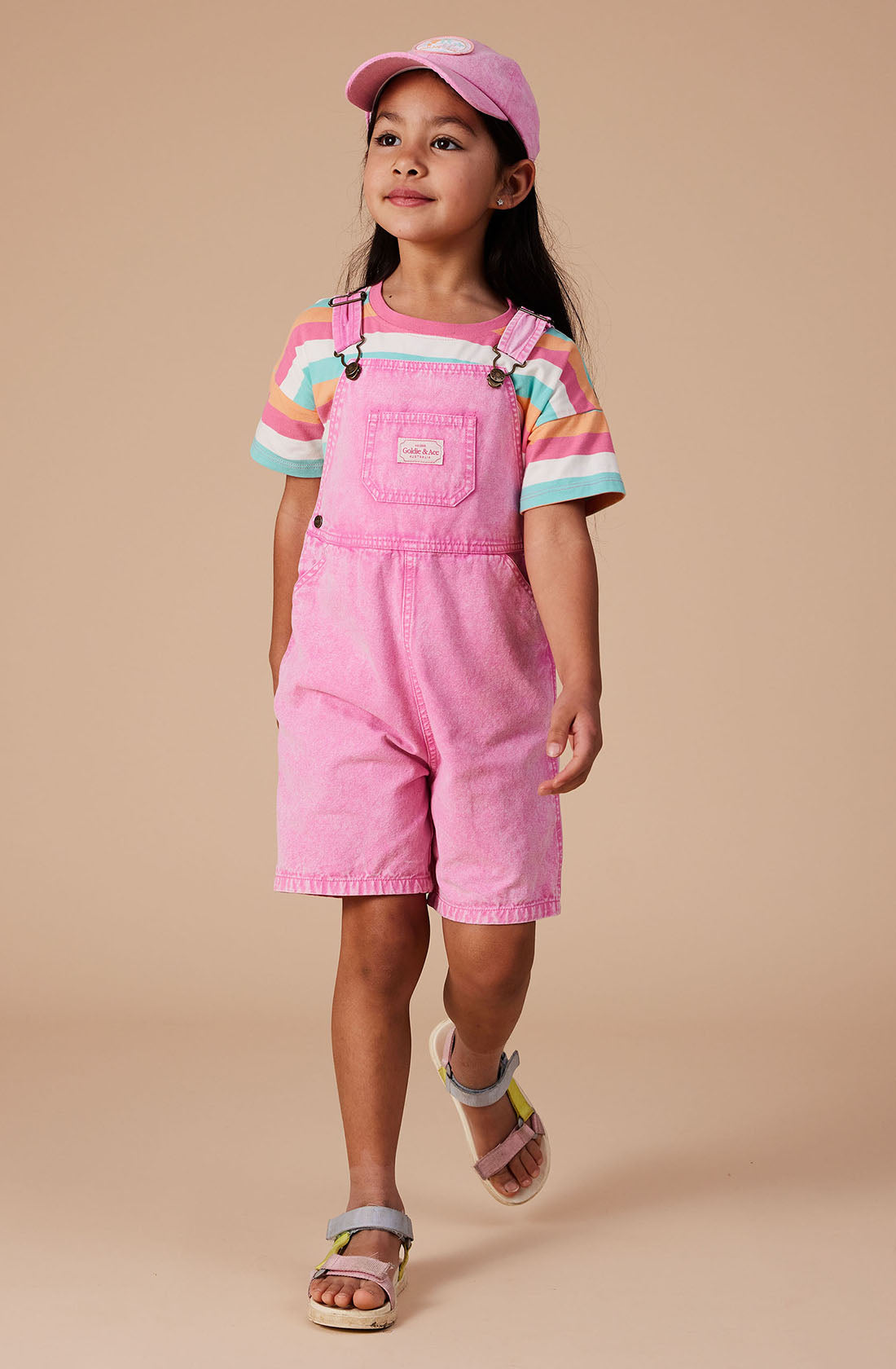 A young girl dons a vibrant pink cap and Burton Vintage Washed Denim Overalls by GOLDIE + ACE, complete with adjustable shoulder straps as she walks confidently. Her long dark hair cascades over a colorful striped t-shirt paired with stylish sandals. The plain, neutral background ensures all attention is drawn to her striking outfit and assured demeanor.